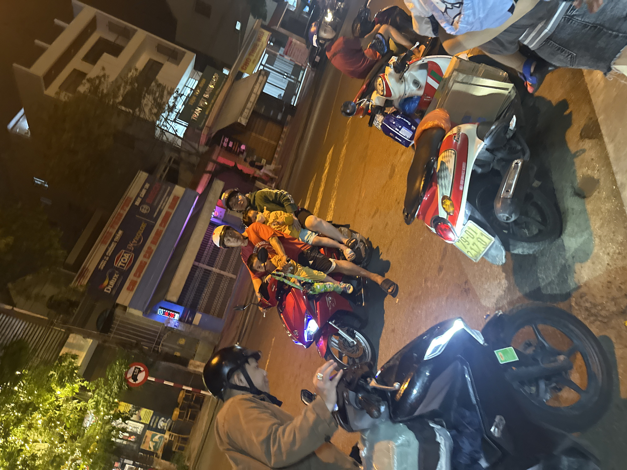 The image, taken at night in Bac Nha Trang Ward, Vietnam, shows a street scene with several parked motorbikes. There are people on the motorbikes and standing nearby. A group of three people is crowded onto one red motorbike, including what appears to be a child. The foreground contains a person on a dark motorbike wearing a helmet and a light-colored jacket. Buildings are visible in the background, including storefronts with signs from businesses such as Dulux, TOA, and Tran. The sign on the motorbike has the registration 79-01 217.01.