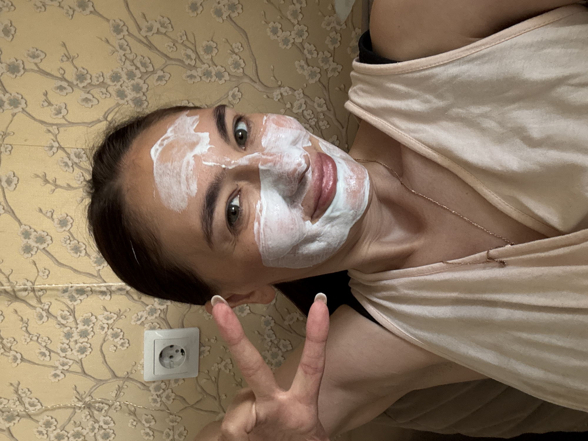 A woman is lying down, angled towards the camera, with a white facial mask applied. She is making a peace sign with her left hand. She is wearing a light pink tank top and a thin gold necklace. The background is a wall with floral patterned wallpaper. A white electrical outlet is visible on the wall near her head. I cannot determine the location is the city of Sochi, Russia, or anywhere else based on this image alone.