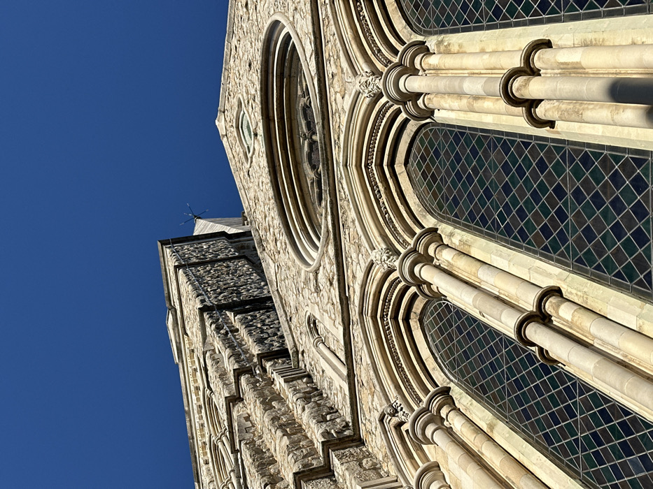 A low-angle photograph captures a detailed section of the exterior of Rochester Cathedral, located in Rochester, Kent, United Kingdom. The structure is primarily composed of light-colored, rough-hewn stone, exhibiting various textures and mortar lines, with smoother dressed stone utilized for decorative elements and architectural features. In the upper-central portion of the image, a large, circular rose window with intricate stone tracery is visible against a clear, deep blue sky. Directly beneath this, three tall, arched windows dominate the lower half of the frame. Each of these windows contains diamond-patterned leaded or stained glass, predominantly in dark green and blue hues. The windows are framed by slender columns and decorative arches, with carved corbels or heads positioned above the apex of the arches. To the left, a receding section of the cathedral displays additional textured stone and stepped architectural lines. A small, spiky metal fixture, possibly a lightning conductor or decorative finial, is discernible on a higher, more distant part of the structure against the sky. No individuals, specific actions, or interactions are present within the scene.