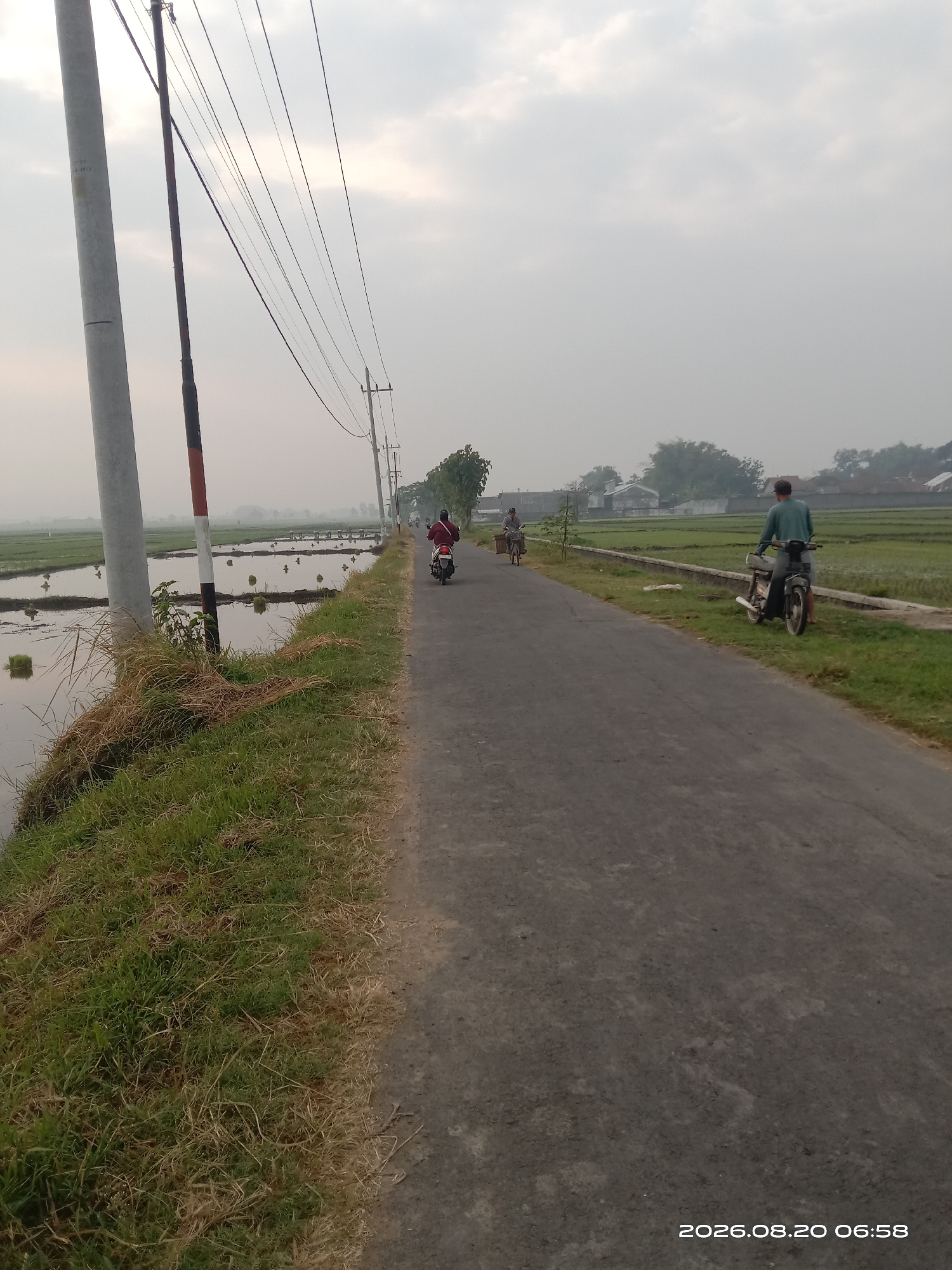 The image depicts a rural road in Bugasur Kedaleman, Indonesia, on a hazy morning. On the left side of the road, there are flooded rice paddies with a utility pole and wires. The road itself is paved and extends into the distance. A man on a motorcycle, wearing a green shirt and a cap, is parked on the right side of the road, facing away from the camera. Further down the road, another person on a motorcycle, wearing a red shirt, is visible. In the background, there are houses and more rice fields. The sky is overcast, suggesting either dawn or dusk, or generally hazy weather conditions. The timestamp at the bottom right indicates August 20, 2026, at 06:58.