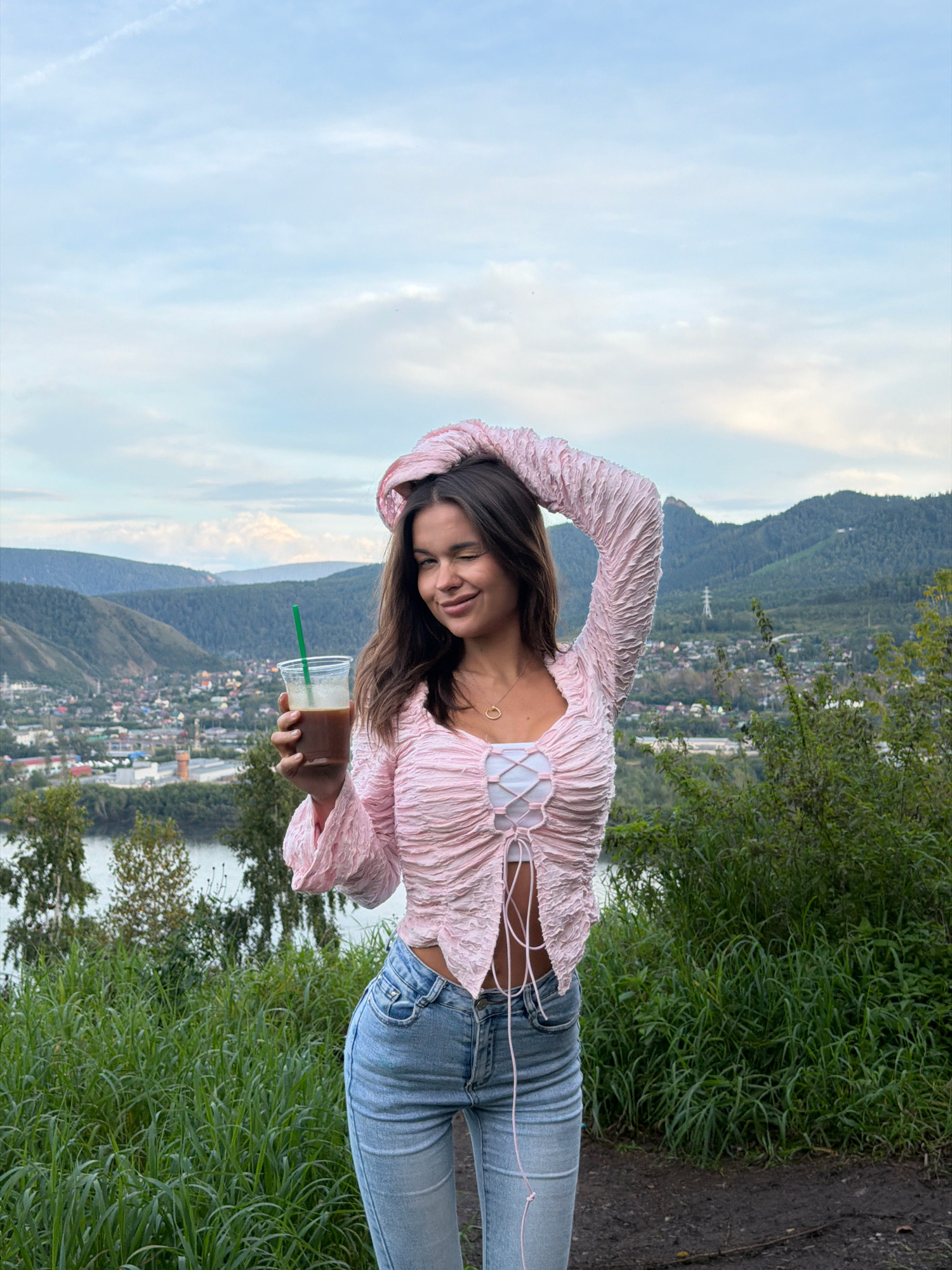 A young woman with long, dark brown hair stands outdoors, holding a clear plastic cup with a green straw filled with a dark beverage. She is wearing a pink, textured long-sleeved top with a tie-front detail and a white underlayer, along with light-wash denim jeans. Her left arm is raised, with her hand resting on her head, and she is winking at the camera. She appears to be standing on a grassy embankment overlooking a city with a river, and mountains in the background. The sky is cloudy and appears to be during the day. The location is identified as Krasnoyarsk, Russia, specifically the viewpoint from the highest point of the National Nature Reserve "Stolby" overlooking the city and the Yenisei River.