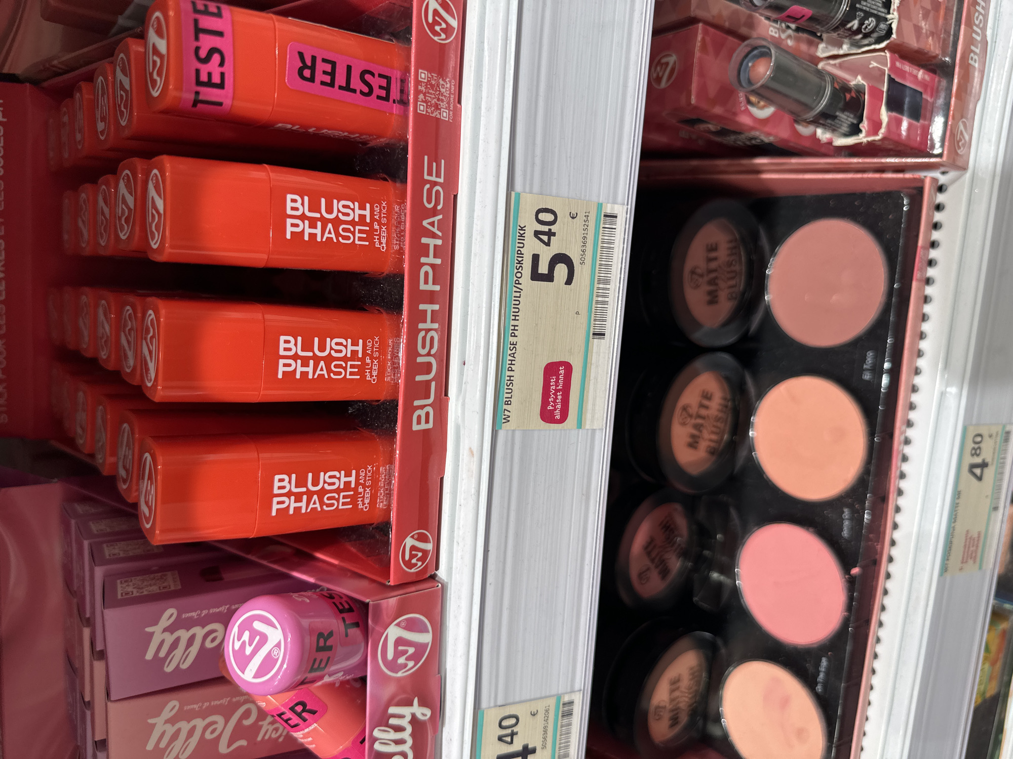 The image is a close-up, eye-level shot of makeup products displayed on store shelves. The dominant colors are pink and orange, with some white and black elements. In the foreground, on the bottom shelf, are several round compacts of blush, some with their lids open showing the powder inside. The compacts are labeled "MATTE BLUSH". Above these, on a middle shelf, are rows of bright orange "BLUSH PHASE" pH lip and cheek sticks. Some of these sticks have "TESTER" labels. The top shelf displays pink packaging for "Jelly" products. A price tag for "W7 BLUSH PHASE PH HUULI/POSKIPUIKK" shows a price of 5.40 €. Another price tag on the bottom shelf for "W7 JOSEPHINE MATTE ME" shows a price of 4.80 €. The products are neatly arranged in a retail display. The environment appears to be a cosmetic store or a section within a larger retail store. There are no people visible in the image.