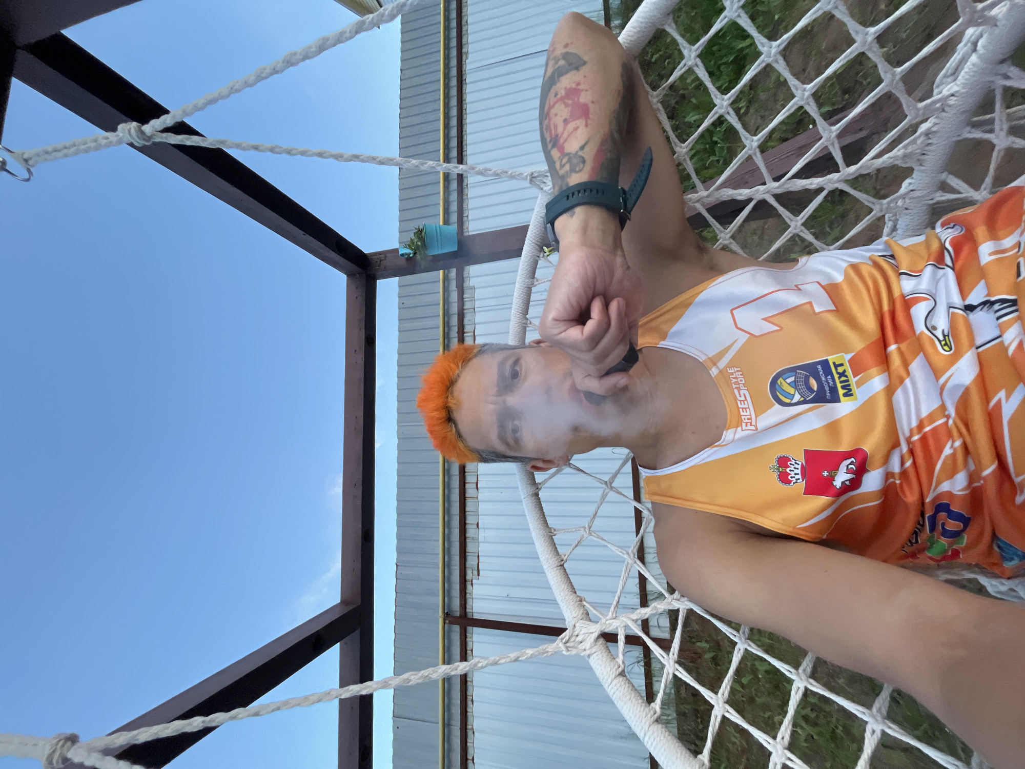 A man with bright orange hair sits in a white hammock chair, exhaling smoke. He is wearing an orange and white athletic tank top with various logos and the number '1' visible. His left arm, which is resting on the armrest of the chair, is covered in tattoos. The hammock is suspended from a wooden frame. In the background, a corrugated metal fence and some greenery are visible. The sky above is a clear blue. This scene appears to be outdoors.