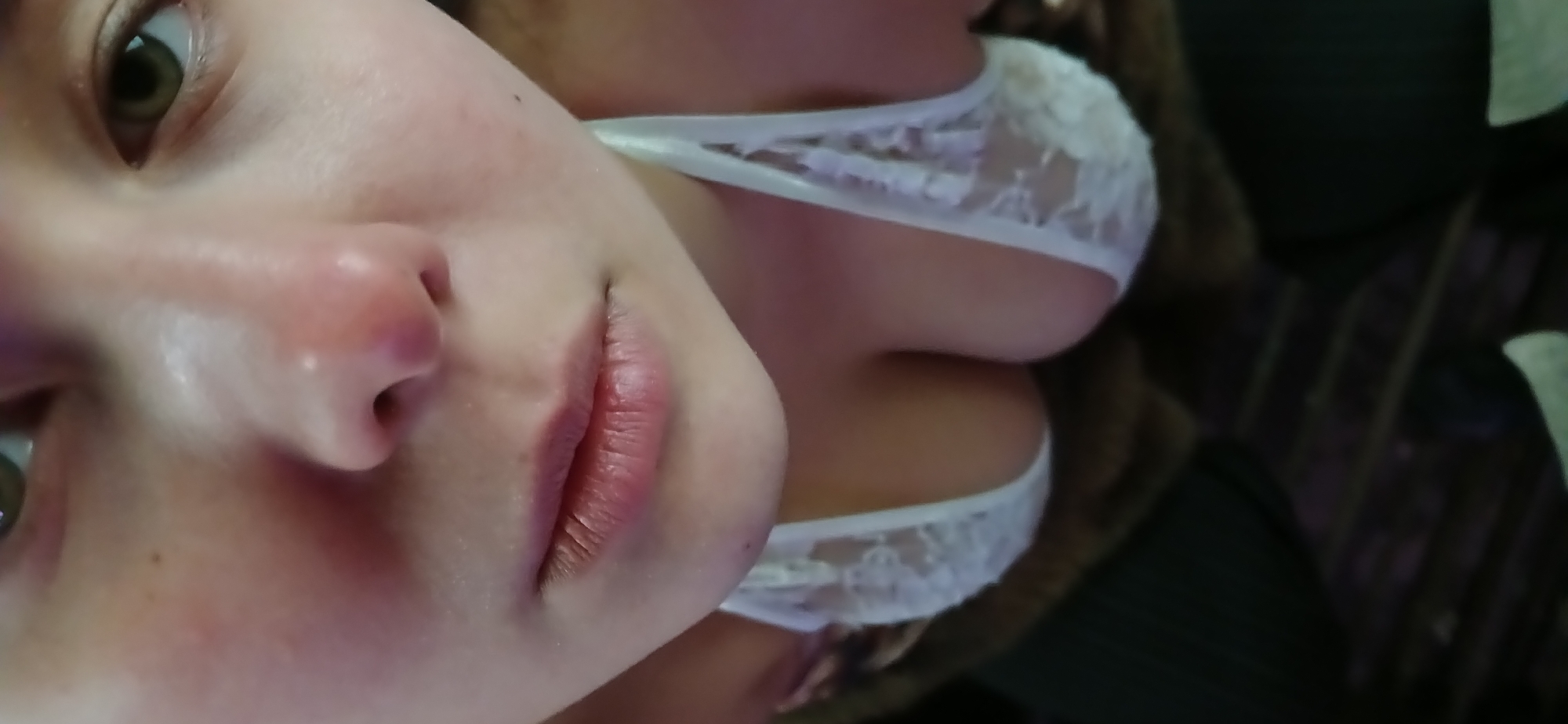 This close-up portrait, contextualized as being from Moscow, Russia, features an individual, likely a female, captured in an intimate indoor setting. The image is significantly rotated, presenting the subject's face and upper body at an oblique angle.
The upper portion of the frame focuses on the subject's face, showcasing a light-colored eye (appearing brown or hazel), the nose, and the upper lip. The skin appears fair, and the lips have a natural, reddish tone. Below the face, a white, lacey undergarment, resembling a bra or bralette, is clearly visible. This garment features delicate floral or geometric lace patterns and is worn on the upper torso, revealing cleavage.
The background is dark and out of focus, providing minimal specific detail about the surroundings beyond indicating an indoor location. The overall impression is a candid, close-up shot, possibly a selfie, emphasizing facial features and a portion of the upper body. The subject appears to be posing for the camera. No specific activities, time of day, weather, or identifiable location cues beyond the provided context are present. There is no visible text in the image.