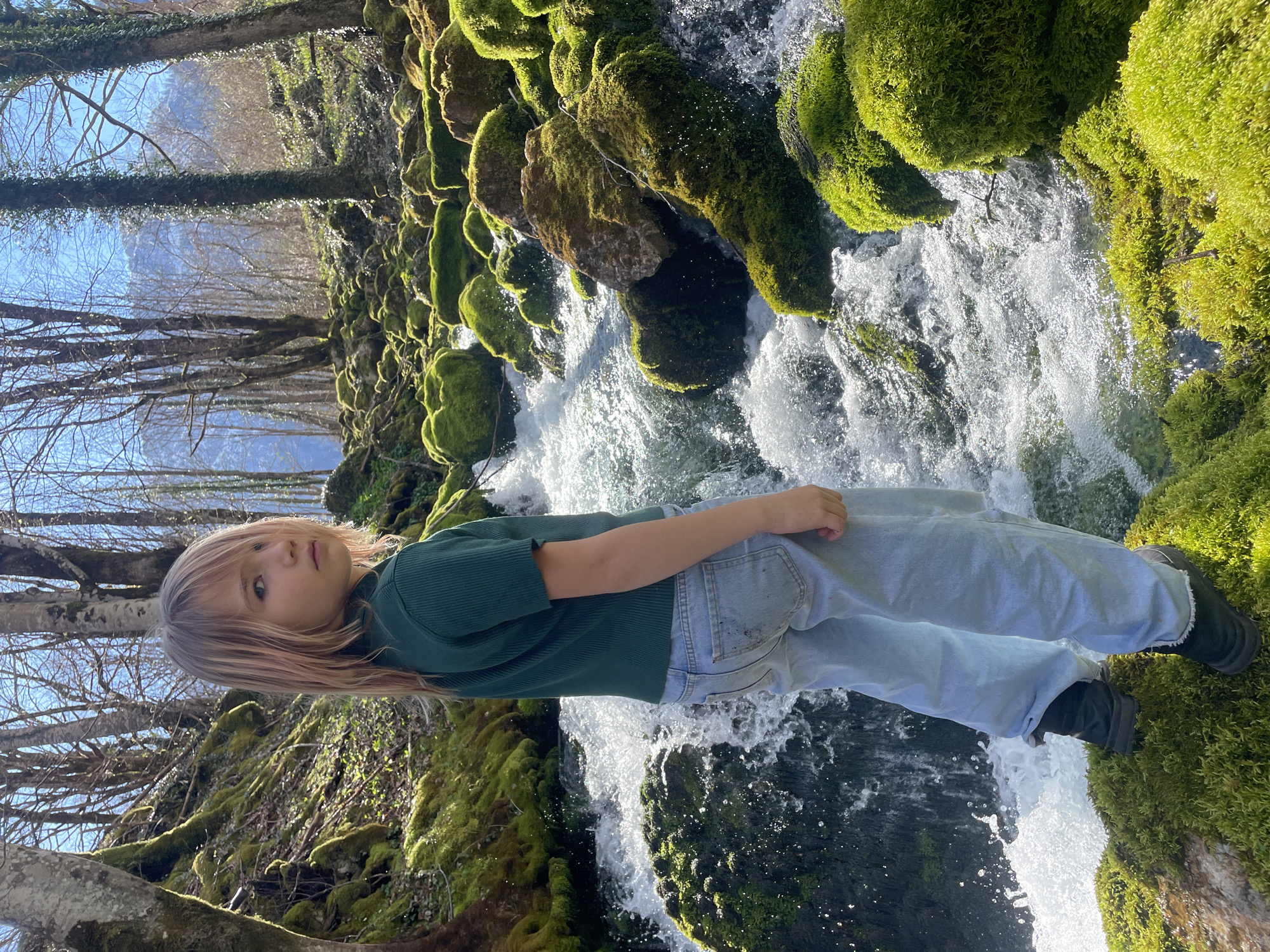 A young person stands on a moss-covered bank beside a waterfall. The person wears a dark green shirt, light blue pants, and dark shoes. Water rushes over rocks covered in moss. Trees are visible in the background under a blue sky. The location is likely near a waterfall in or around Bistrica, Montenegro.