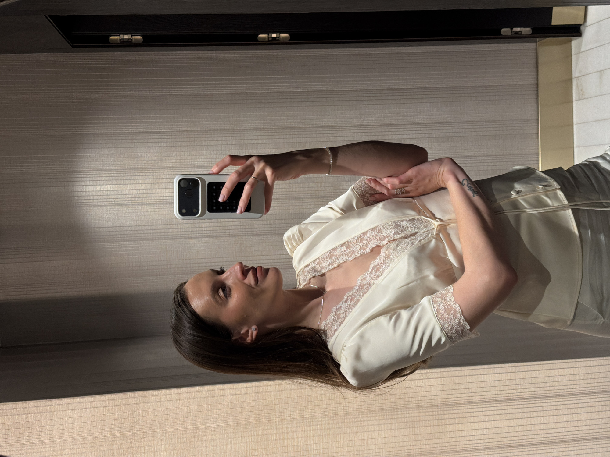 A woman with long brown hair and wearing a cream-colored silk garment with lace trim is holding up a smartphone and taking a selfie in an interior setting, likely a hotel room. Her left arm is extended to hold the phone, and her right hand rests near her chest. She has a bracelet on her left wrist and a tattoo on her right forearm. The background consists of a wall with horizontal stripes and a dark cabinet above. The setting appears to be indoors in Budva, Montenegro, possibly a hotel room.