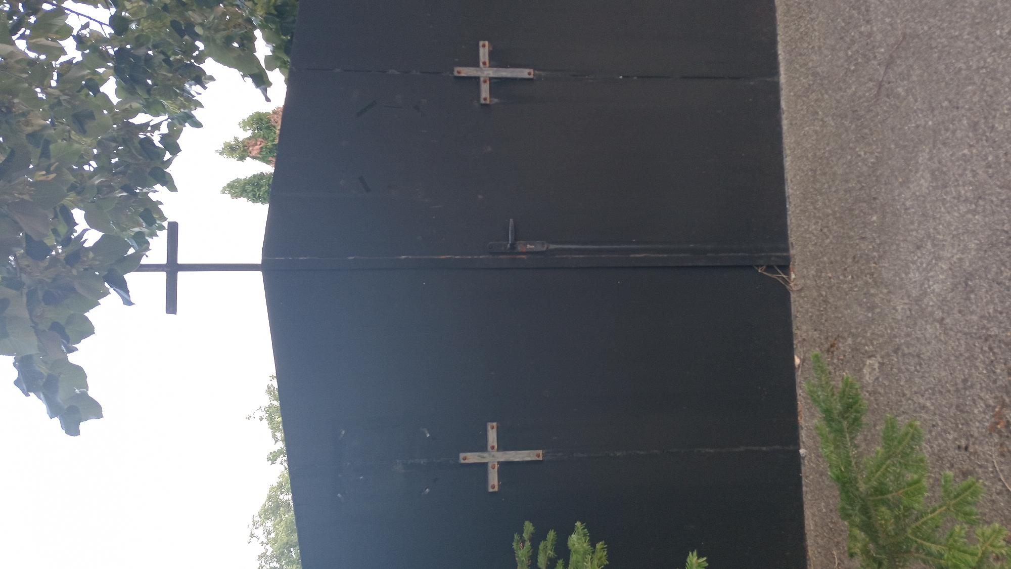 In the city of Dolno Izvorovo, Bulgaria, a dark metal gate is visible, featuring two imposing doors. Mounted centrally at the top of the gate is a large, dark cross. On the left door, a smaller, metallic cross is affixed, with visible rivets. Similarly, on the right door, another metallic cross is attached, also with apparent rivets. A black door handle is positioned on the right door. The gate is bordered by a grey, gravel-like surface, with a sprig of green foliage in the lower left corner. Above the gate, the tops of trees and branches are discernible against a bright, overcast sky.