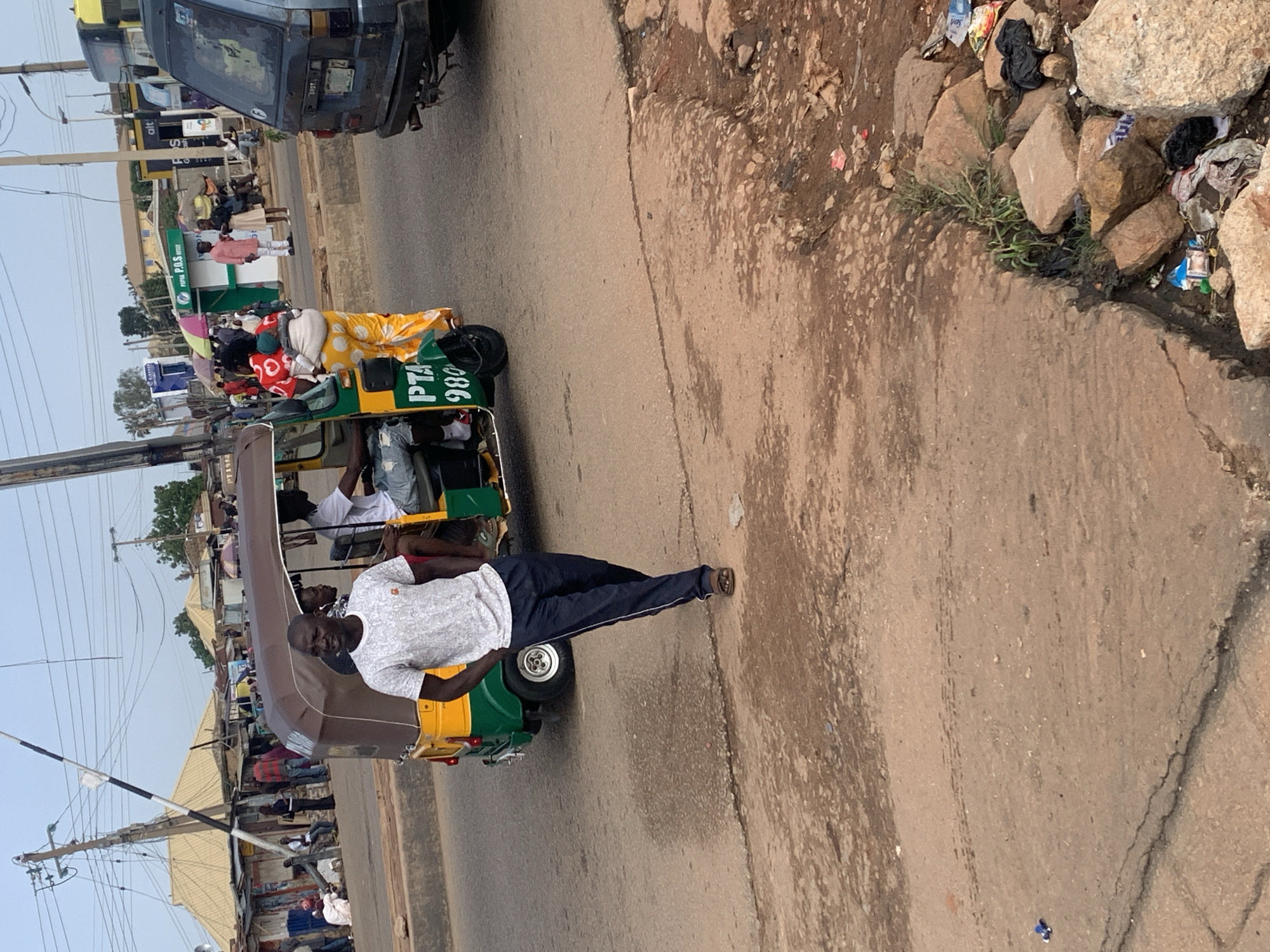 In Gura Top, Nigeria, a street scene unfolds with a focus on transportation and pedestrian activity. A man in a grey t-shirt and dark pants walks on a dirt and asphalt road, his hands in his pockets. Behind him, a yellow and green auto-rickshaw, a common mode of transport, is partially visible, with two passengers inside. The auto-rickshaw has the letters "PTA" and the number "980" displayed on its side.
The background reveals a bustling commercial area with various structures, including shops with corrugated metal roofs and awnings. People are seen walking and gathered, suggesting a vibrant marketplace or public thoroughfare. A utility pole with wires stretches across the scene, indicating the presence of infrastructure. Further in the distance, more buildings and vegetation are visible, hinting at the broader urban landscape. To the right of the auto-rickshaw, a dark blue van is parked. The ground is a mix of dirt, gravel, and patches of asphalt, with some areas showing signs of wear and tear. Litter and debris are scattered along the roadside, particularly in the lower right portion of the image, where rocks and trash are accumulated. The lighting suggests it is daytime.