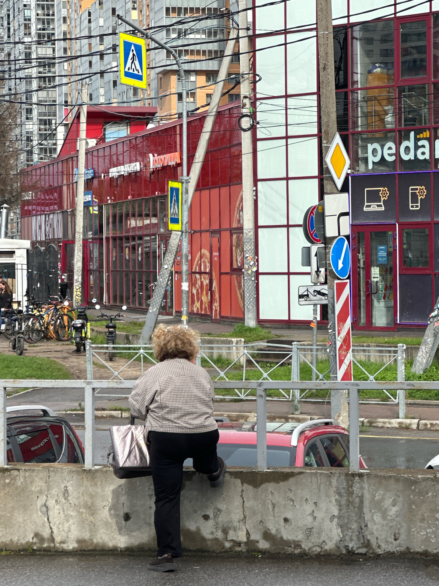 In Murino, Russia, a woman with curly blonde hair is perched on a concrete barrier, facing away from the camera and holding a metallic shopper bag. She is wearing a checkered shirt and black pants. In the background, a red building with large glass windows and architectural details stands prominently. Several bicycles and scooters are parked to the left of the building. A pedestrian crossing sign is visible on a pole above the street, and further down, a cluster of traffic signs is mounted on a pole. Two red cars are partially visible beyond the concrete barrier. The scene is set on a city street with buildings and power lines extending into the distance.