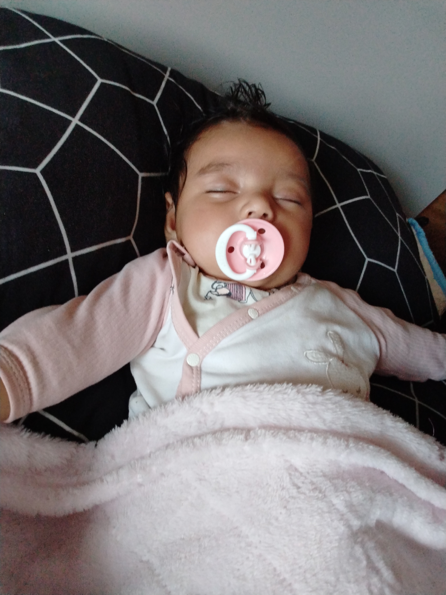 A sleeping baby is lying on a black and white patterned pillow. The baby has dark hair and is wearing a white and pink onesie. A pink pacifier is in the baby's mouth, and a pink blanket covers the baby's torso. The location is in the city of Unknown, Unknown.