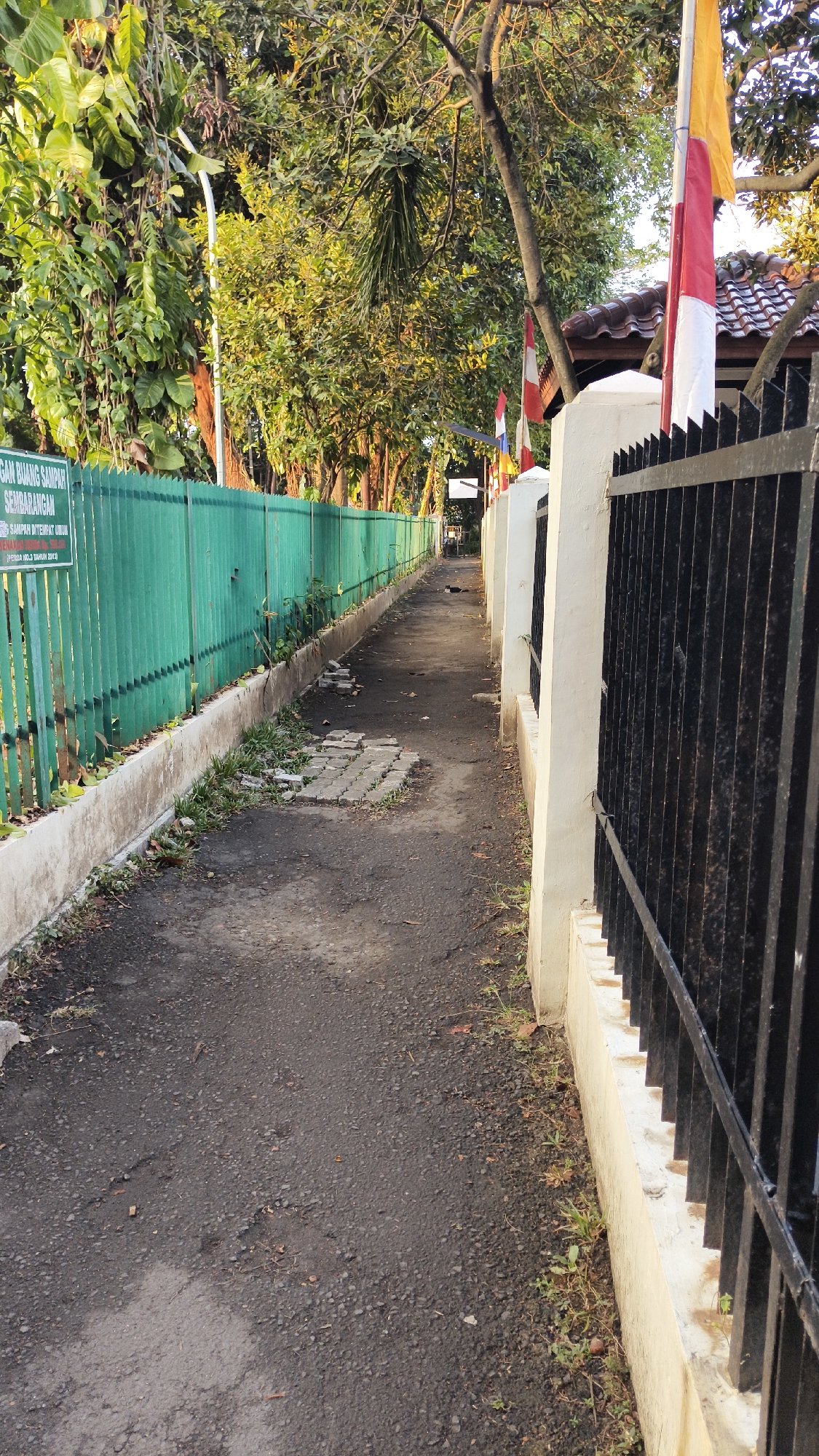 This is a photograph of a paved pathway leading away from the viewer, bordered by a green metal fence on the left and a black metal fence with white concrete pillars on the right. The pathway is asphalt, with some cracks and patches. On the left side, the green fence has a sign with Indonesian text that reads "DILARANG BUANG SAMPAH SEMBARANGAN" (No littering). Vegetation, including grass and weeds, grows along the base of both fences. Trees with lush green foliage are visible behind the fences, illuminated by sunlight. Two Indonesian flags, red and white, are visible flying on poles above the black fence on the right. The setting appears to be a public space, potentially a park or a walkway in a residential or institutional area within the city of South Jakarta, Indonesia. The lighting suggests it is daytime, possibly late afternoon due to the warm, golden hue of the sunlight. There are no people or animals clearly visible in the scene, though a small dark object that might be a bird is present further down the pathway on the right side.