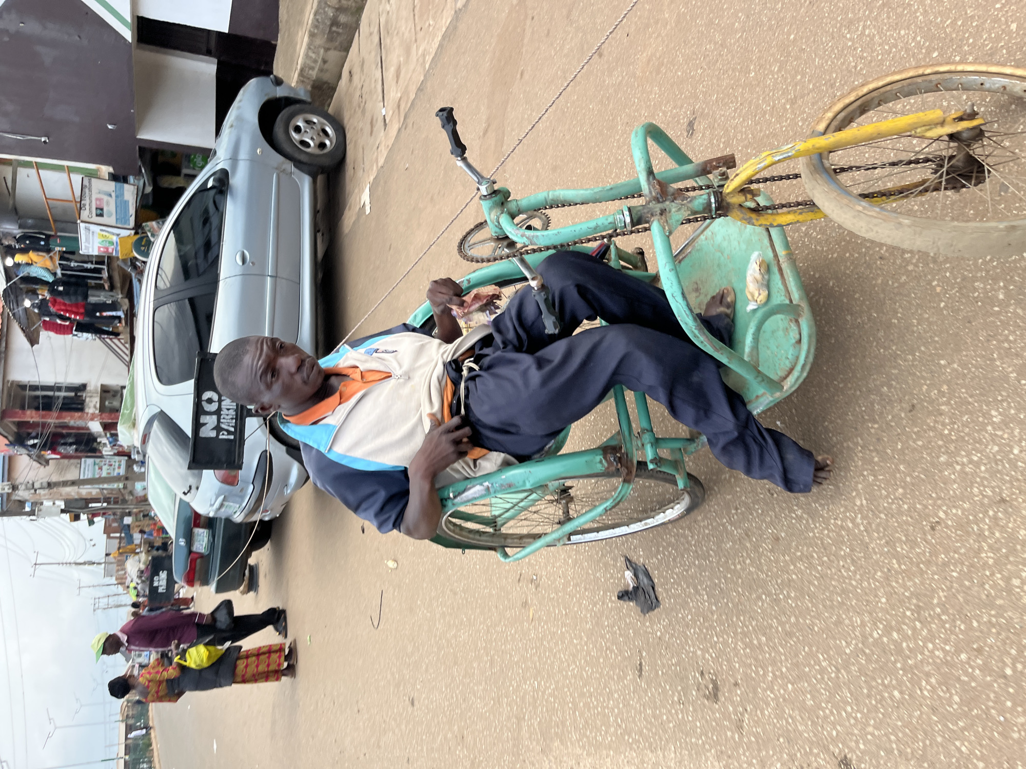 A man in a wheelchair is seen from a medium shot, looking at the camera. He is wearing a blue, white, and orange jacket over an orange shirt, and dark blue pants. The wheelchair is a three-wheeled vehicle, painted green, with two large rear wheels and a smaller front wheel. The man's hands are on the controls of the wheelchair. The wheelchair is on a paved street with scattered debris. In the background, there are cars, buildings, and other people. Specifically, a silver car with a "NO PARKING" sign is visible. Further in the background, other vehicles, streetlights, and shops can be seen, indicating an urban street scene. The overall environment suggests a busy street in Bukuru, Nigeria.