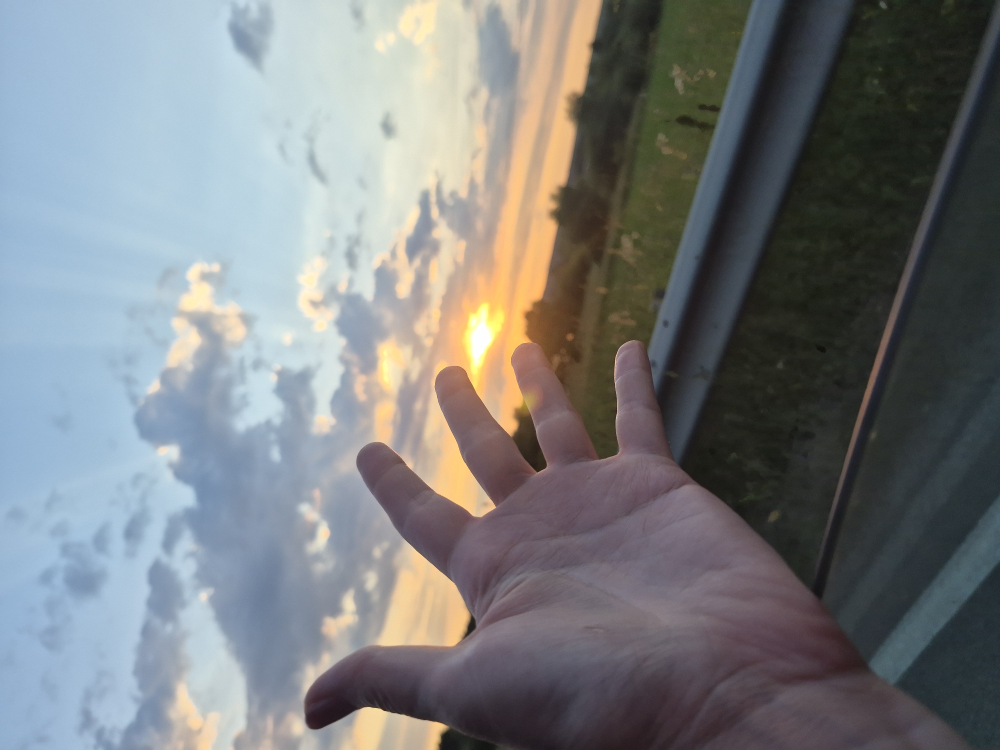 The image is a close-up shot of a person's open left hand reaching out towards a sunset. The hand, with fingers spread, is in the foreground and slightly out of focus. Behind the hand, the sky is filled with dramatic clouds, some illuminated by the setting sun, creating crepuscular rays. The sun itself is a bright orb near the horizon, casting a warm orange and yellow glow. Below the sky, there is a landscape of green fields and trees, with a road guardrail visible in the middle ground, separating the road from the field. The scene appears to be captured from a vehicle, given the perspective and the guardrail. The location is stated to be Ol'khovo, Russia.