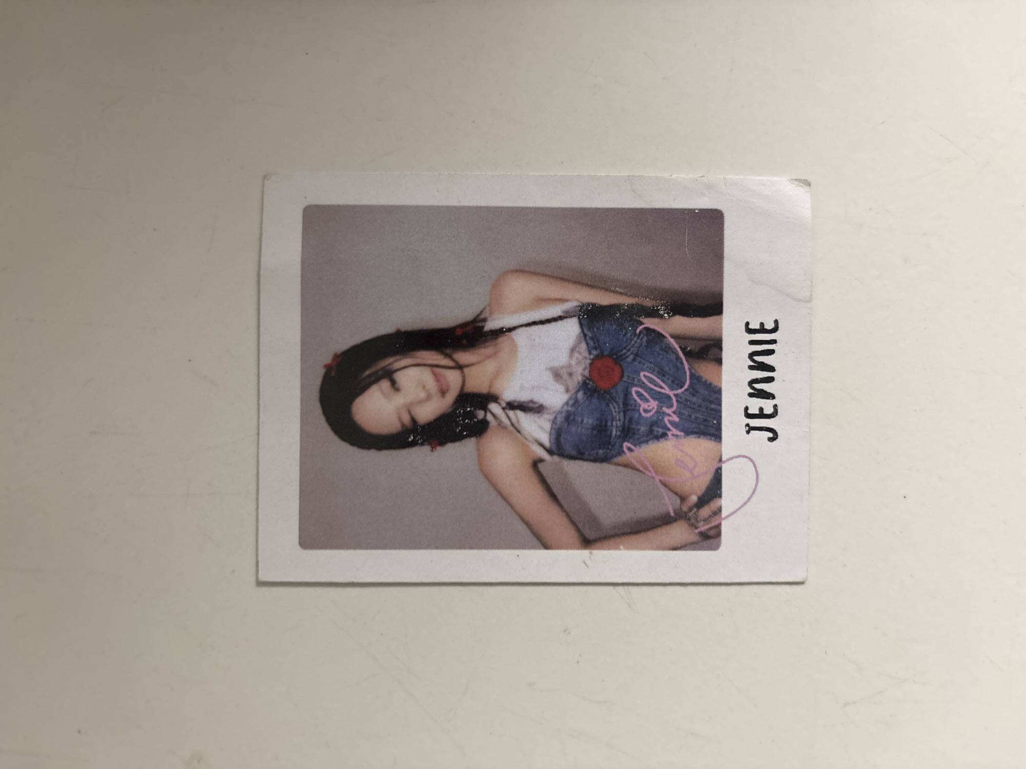 A photograph, presented in a white border, depicts a young woman with dark hair styled in braids, wearing a denim bustier over a white top. She is posing with her right hand on her hip, her left arm bent with her hand on her chest, and her head tilted slightly to the side. A pink signature is visible on her chest, and the name "JENNIE" is printed in black text below the photograph. The background is a plain, light gray. The photograph is situated on a light-colored, textured surface that appears to be a wall or tabletop. There are no identifiable landmarks or elements that would indicate a specific location in North Jakarta, Indonesia.