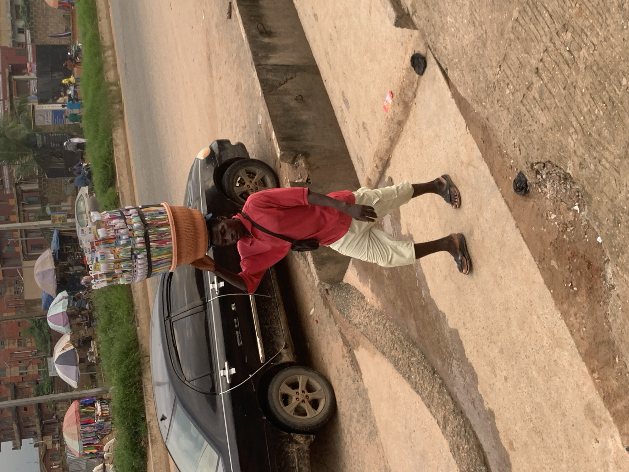 A man is walking on a dirt and concrete path next to a black car. He is wearing a red shirt and light-colored pants, and he is carrying a large basket of toothbrushes on his head. The basket is round and made of woven material. In the background, there are buildings, trees, and other people. The scene appears to be a street in Shagamu, Nigeria.