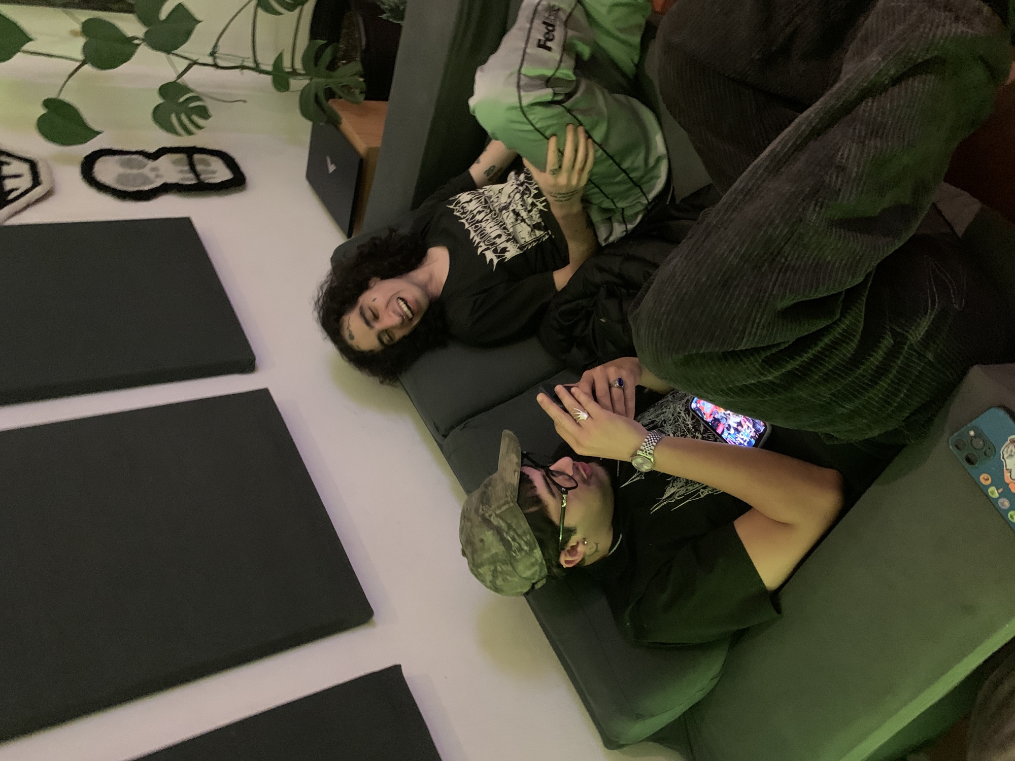 Two young men are reclining on a dark gray sofa. One is smiling and facing the camera, wearing a black shirt with a white design and gray sweatpants with green stripes. The other is wearing a camouflage baseball cap and looking at a cell phone. A watch is on the wrist closest to the camera and there is a blue cell phone lying next to him. There is a monstera plant to the left of the sofa. Black and white decorations are on the wall. There are square, black acoustic panels on the wall. The location cannot be determined from the image.