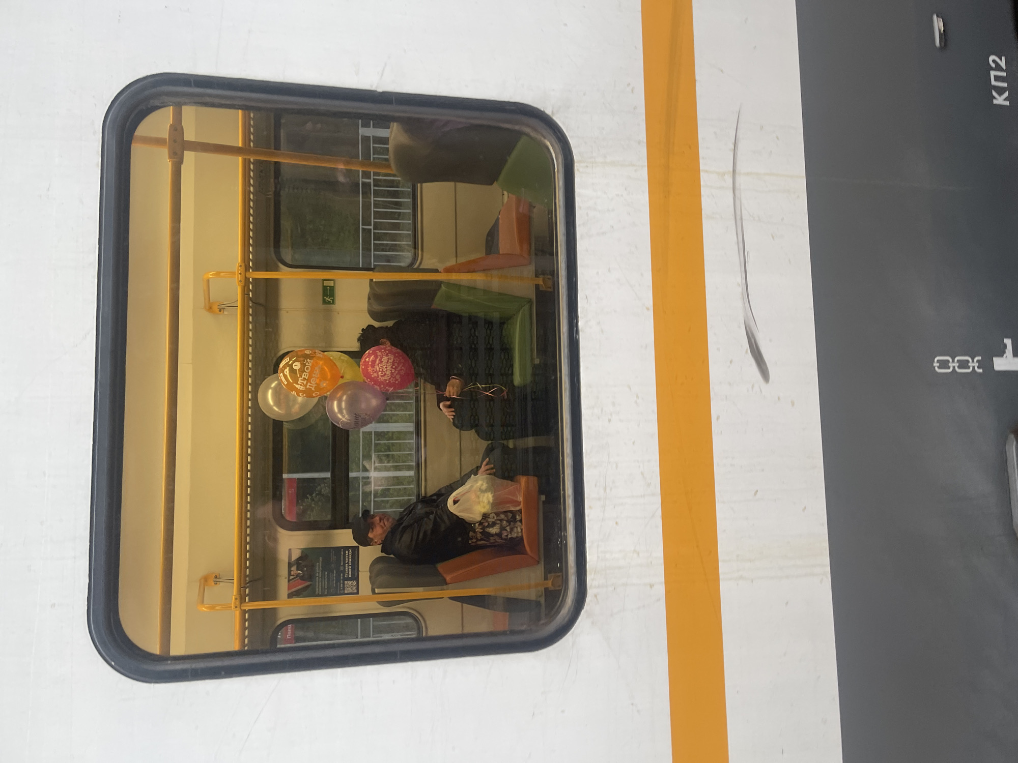 The image shows a rectangular window of a train carriage. Inside the carriage, there are people sitting, and a bunch of balloons. Outside the train, there's white and grey paint with an orange stripe. The Cyrillic inscription "КП2" is visible. The location could possibly be Trudovaya, Russia.