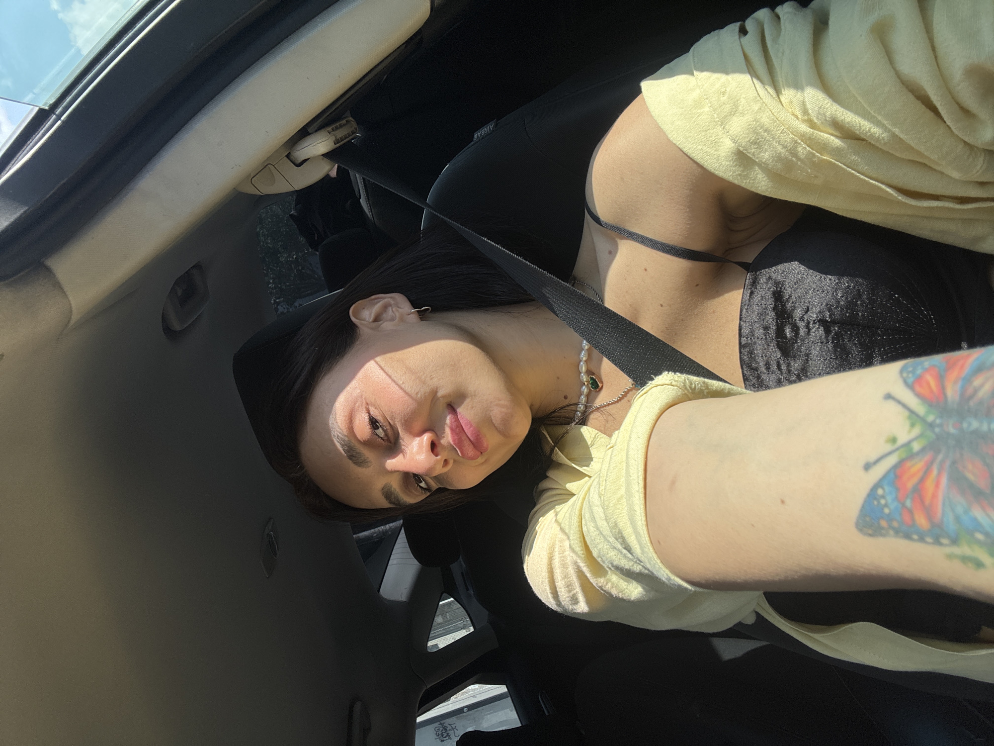 A young woman with dark hair and light skin is taking a selfie while sitting in the driver's seat of a car. Sunlight streams in from the right, illuminating her face and casting shadows. She is wearing a black camisole, a light yellow open shirt, and a pearl necklace with a green pendant. Her left arm, adorned with a colorful butterfly tattoo, is extended as she holds the phone. A black seatbelt is fastened across her chest. The background shows the interior of the car, including the dashboard, a window, and part of the passenger seat. The context of Tbilisi, Georgia is not visually apparent from the image.