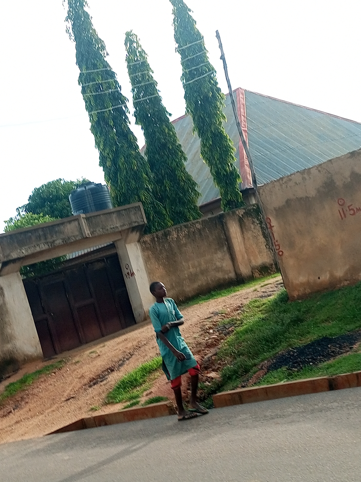 A young boy wearing a teal robe and red shorts stands on a paved street in front of a concrete wall and a large metal gate. He is looking to his right, with his left arm bent and resting on his waist. Behind him, a dirt path leads to the gate, which is flanked by concrete pillars. A black water tank is visible on top of the wall to the left of the gate. Several tall, slender trees with dense green foliage stand to the left of the wall and gate. A building with a gray, sloped roof and red trim is visible behind the trees and to the right of the gate. Red markings, including what appears to be "11.5m," are present on the concrete wall to the right. The environment appears to be an outdoor scene in Tempure, Nigeria, with a mix of urban structures and vegetation.