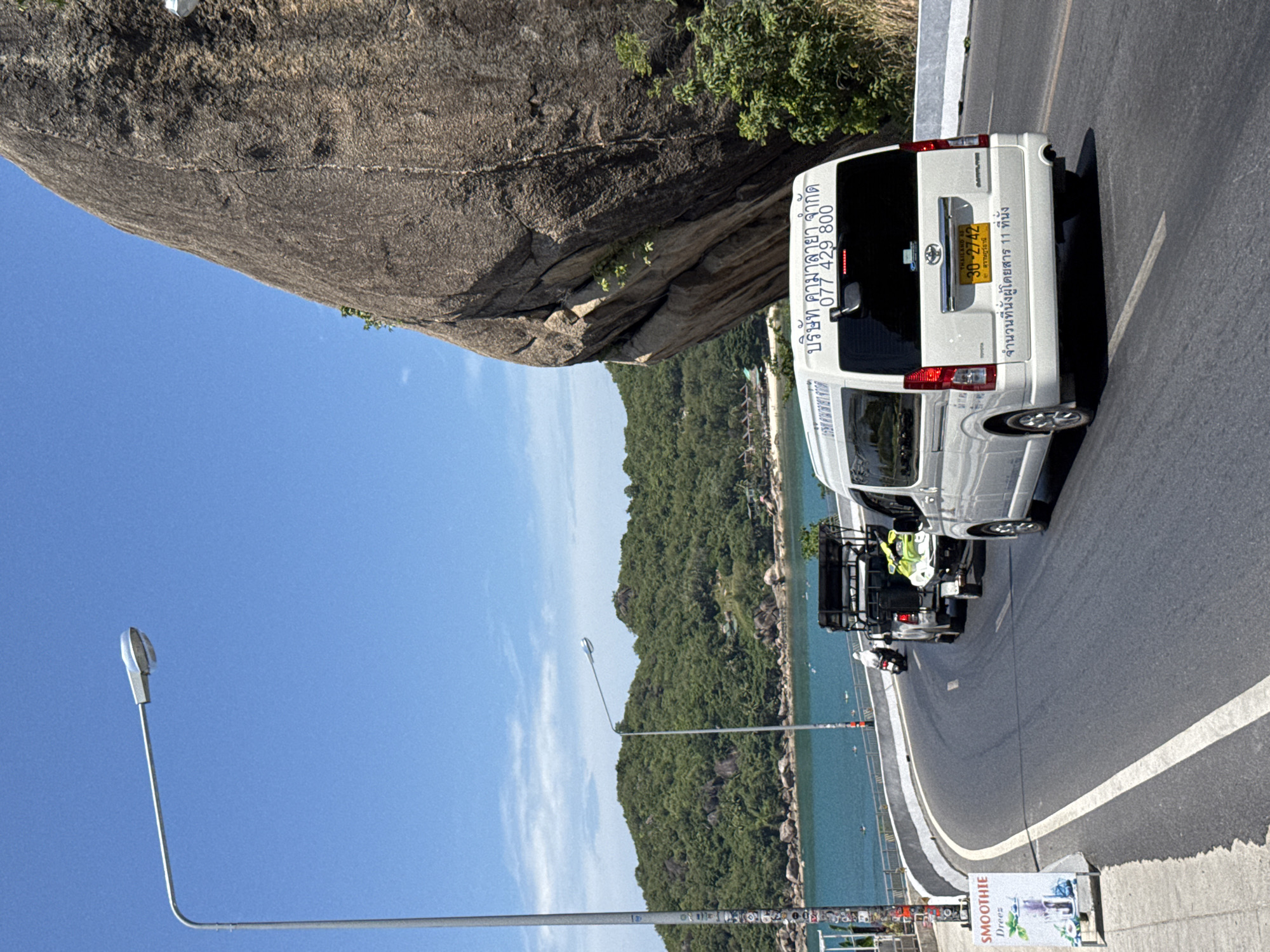 In Ko Samui District, Thailand, on a sunny day, a white van with the license plate 30-2742 is driving on a curved road next to a large rock formation. The van has Thai writing on its rear, indicating it's a transport company with the phone number 077 429 800. A trailer carrying a jet ski is attached to a dark-colored truck that is following the van. A person wearing a white outfit is riding a motorcycle in front of the truck. On the left side of the road, a sign advertises "SMOOTHIE Dreez" with a picture of a beverage. In the background, lush green hills and blue water with small boats are visible. A street lamp stands to the left of the road.