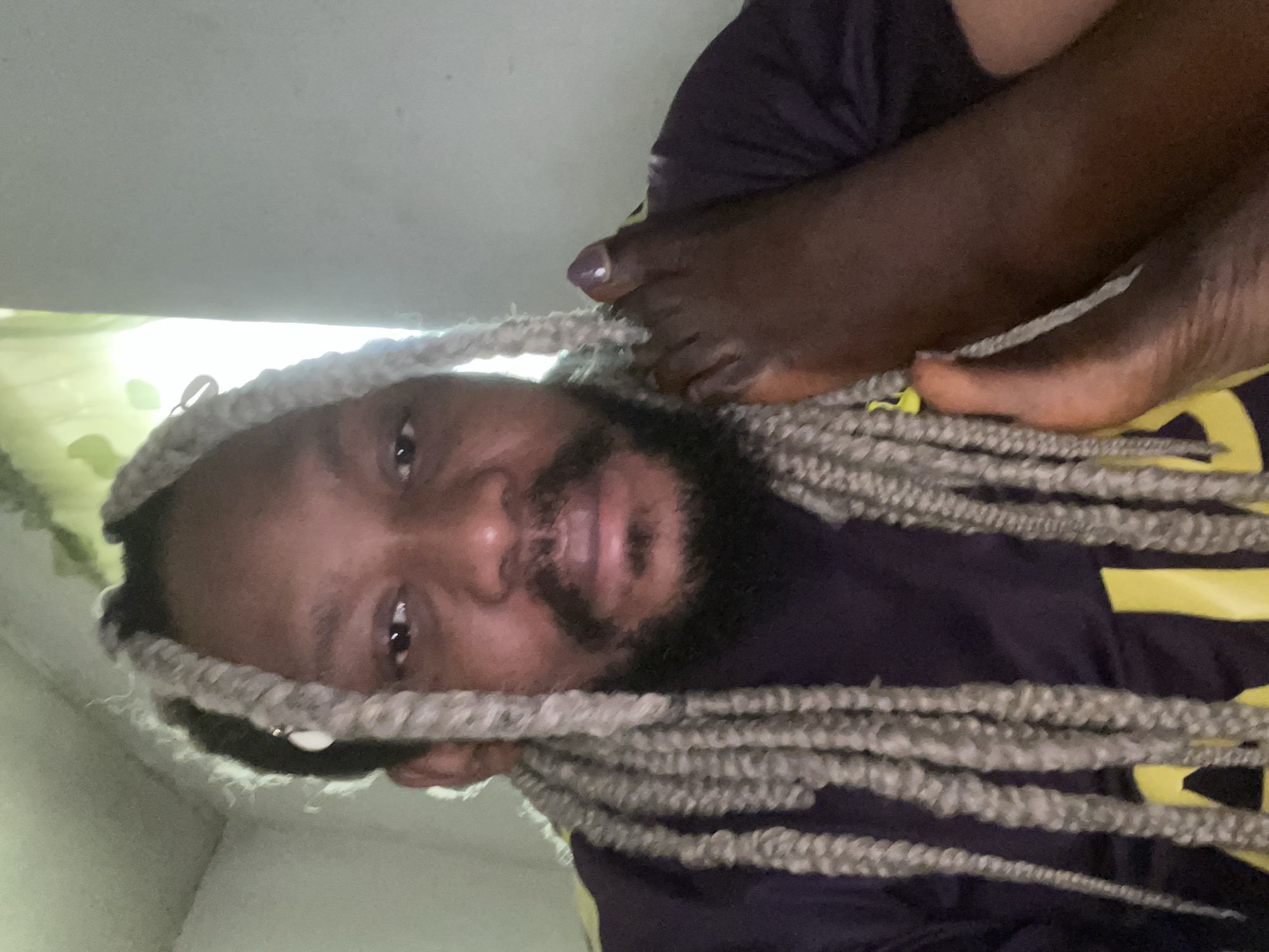 The image shows a close-up of a person with light gray braided hair and a beard. The person is wearing a dark purple t-shirt with yellow accents. Their right foot is raised and resting near their face, with a toenail painted a muted purple color. The background appears to be an indoor setting with a light-colored wall and a window with a green curtain or plant visible. The lighting suggests it may be daytime. The scene does not provide any specific visual cues to pinpoint its location within the city of Jos, Nigeria.
