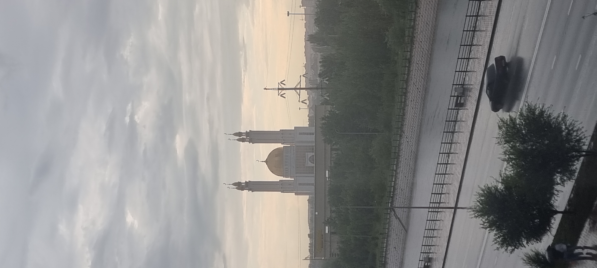 The image captures a panoramic view of the city of Aktobe, Kazakhstan, under an overcast sky, possibly during or after rainfall. In the distance, a prominent mosque with a golden dome and two tall minarets stands as the central architectural feature. The mosque is flanked by lower structures and appears to be situated on a raised platform or hill. Below the mosque, a dense line of green trees forms a natural barrier.
In the foreground, a wet asphalt road is visible, with reflections indicating recent rain. A dark-colored car is captured in motion on the road, its headlights on. To the left of the road, a sidewalk area is lined with trees, and two figures, one possibly holding an umbrella, are walking. The road runs alongside a body of water, likely a canal or river, with a retaining wall and a metal fence separating it from the road. Overhead, power lines and utility poles are visible against the cloudy sky. The overall scene suggests an urban environment with a significant religious landmark and signs of recent precipitation.