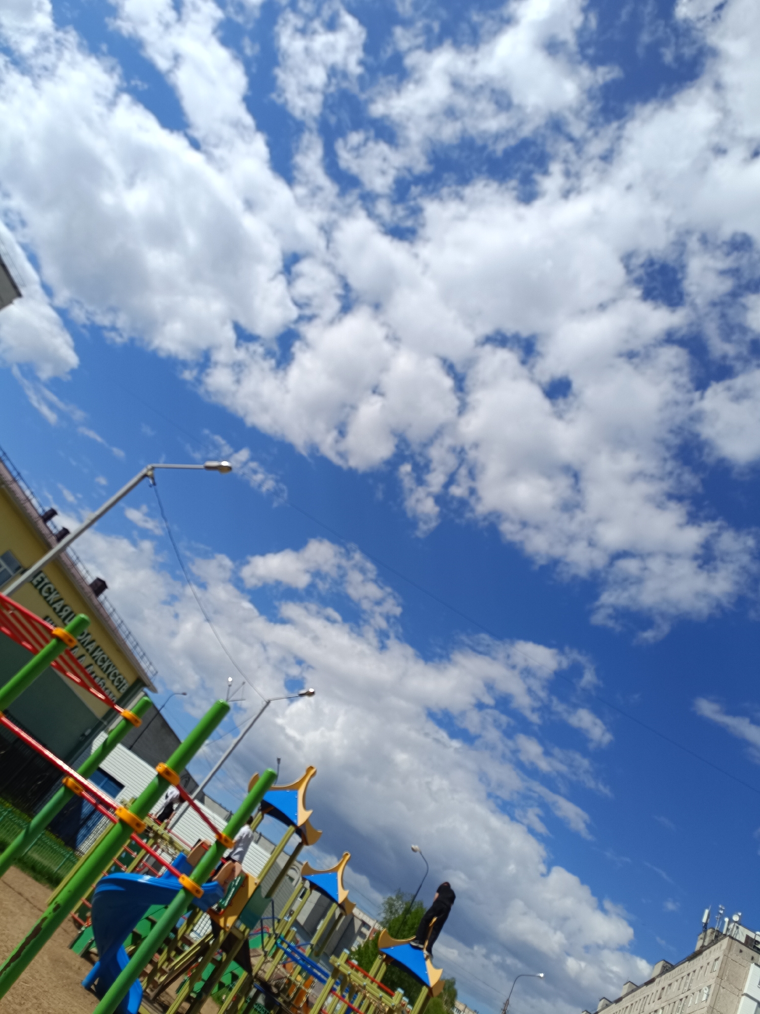 The image shows a playground in Tobolsk, Russia, under a blue sky with scattered cumulus clouds. In the foreground, there is a children's climbing structure with red, green, blue and yellow bars. Behind the playground, there are two multi-story buildings and a street lamp. One person is standing on one of the buildings' roofs.