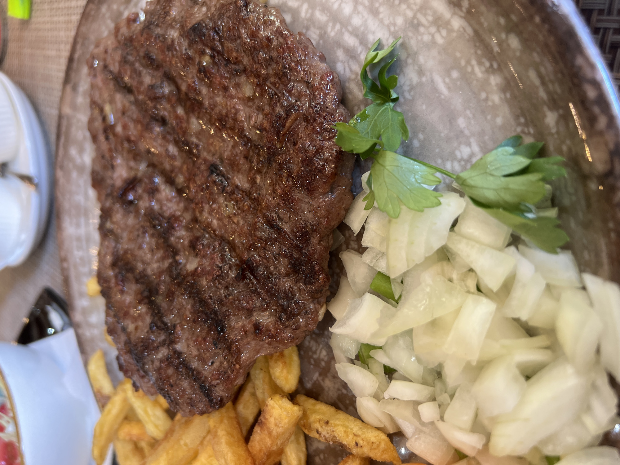 This close-up, overhead shot displays a plate of food, likely from a restaurant in Budva, Montenegro. The primary subject is a grilled meat patty, possibly ćevapi given the context, which is dark brown with prominent grill marks. The patty is glistening, suggesting it's freshly cooked and juicy.
To the right of the meat, a generous portion of chopped white onion is visible, alongside a few sprigs of fresh green parsley. The onions appear crisp and raw, offering a sharp contrast to the savory meat. Below and to the left of the patty are golden-brown french fries, arranged in a somewhat scattered manner.
The food is served on a plate with a textured, mottled brown and grey surface, typical of restaurant dinnerware. The overall lighting suggests an indoor setting, possibly a restaurant dining area during daytime, as there are no strong shadows or indications of natural light. The focus is sharp on the food, with the background slightly blurred, emphasizing the meal itself. There are no people or text visible in the image. The scene is focused solely on the presentation of this savory meal.