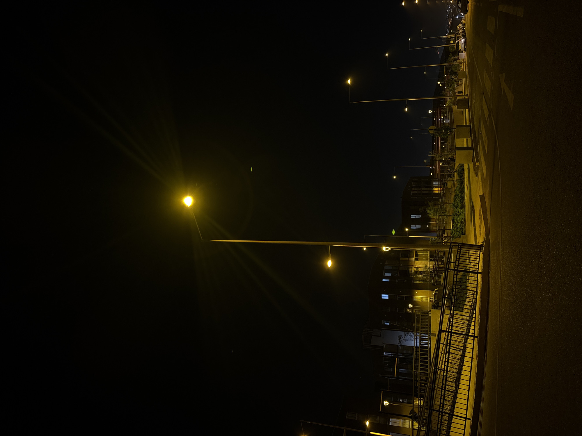 The image depicts a night scene in Arica, Chile. A row of streetlights illuminates a paved road with pedestrian crossings. The scene is dark, with the lights creating strong contrasts. On the left of the road, there is a walkway with a railing. In the background, buildings with illuminated windows can be seen. The overall impression is of a quiet, urban environment at night.