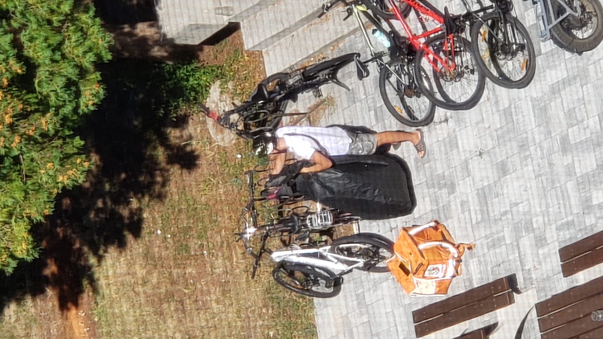 The image shows a street scene in Prešov, Slovakia, with several bicycles parked at a bike rack. A person wearing a white t-shirt, camouflage shorts, and a helmet is holding a large black bag. An orange delivery bag is placed on the ground near a wooden bench. The environment appears to be an outdoor area with trees and paved ground. The lighting suggests it is daytime.
