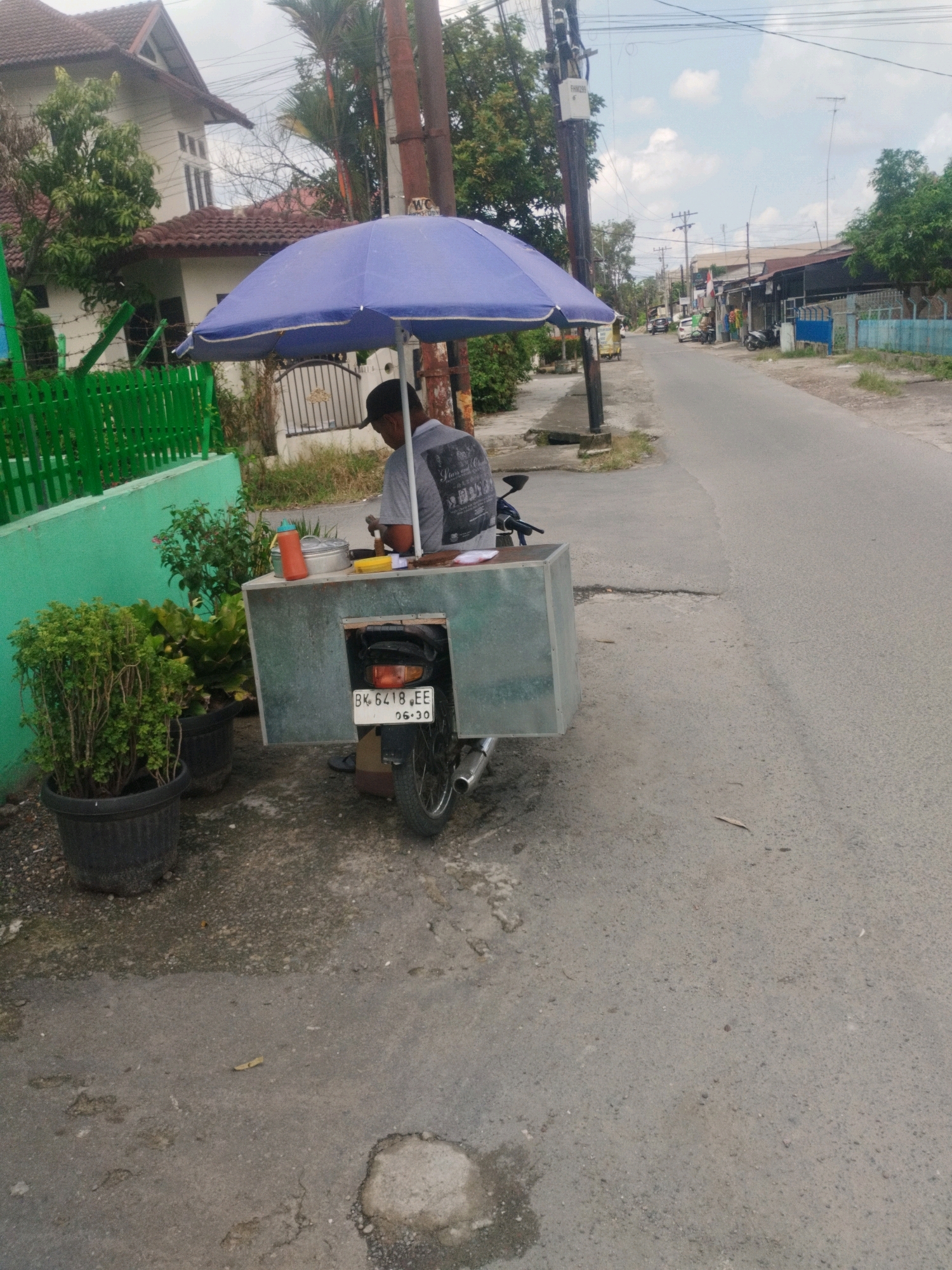 A person is sitting under a blue umbrella next to a food cart mounted on a motorcycle on the side of a paved street in Medan, Indonesia. The person is wearing a grey t-shirt and a black cap and appears to be preparing food. The food cart is made of metal and has a license plate that reads "BK 6418 EE". There are two potted plants in the foreground, one with lush green foliage and another with smaller green leaves. In the background, there is a white house with a red tiled roof, a green fence, and trees. The street is lined with buildings and more trees. The sky is cloudy.