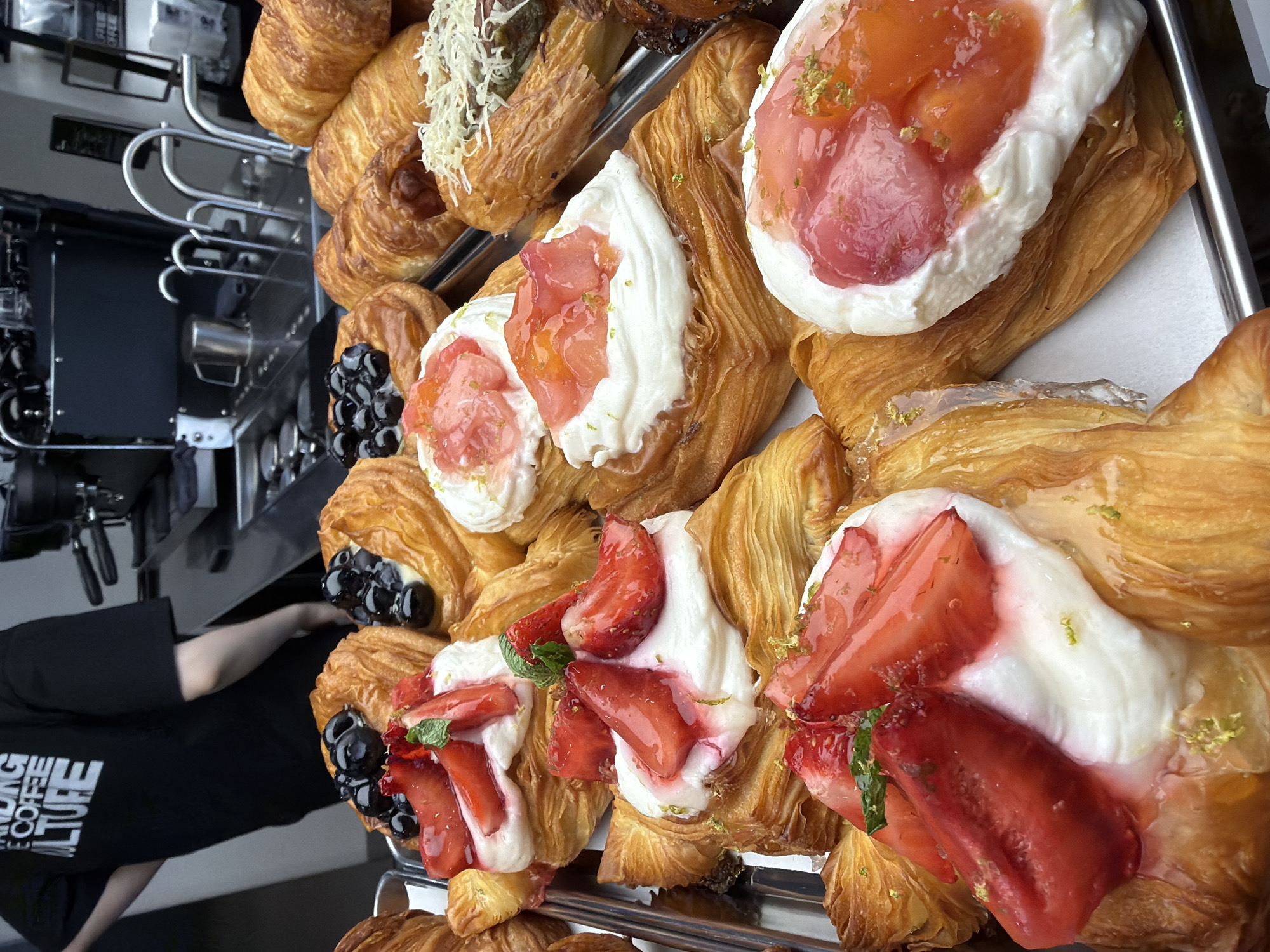 The image shows a close-up of a display of pastries on a metal tray. The pastries appear to be a variety of croissants and danishes, some topped with cream and fruit such as strawberries and blueberries, and others with what looks like grated cheese. In the background, partially visible, is a person wearing a black t-shirt with white text that reads "STANDING COFFEE CULTURE." Behind the person and the pastries, a coffee machine and counter are visible. The scene appears to be set in a cafe. There is no information in the image to suggest the location is in Izhevsk, Russia.