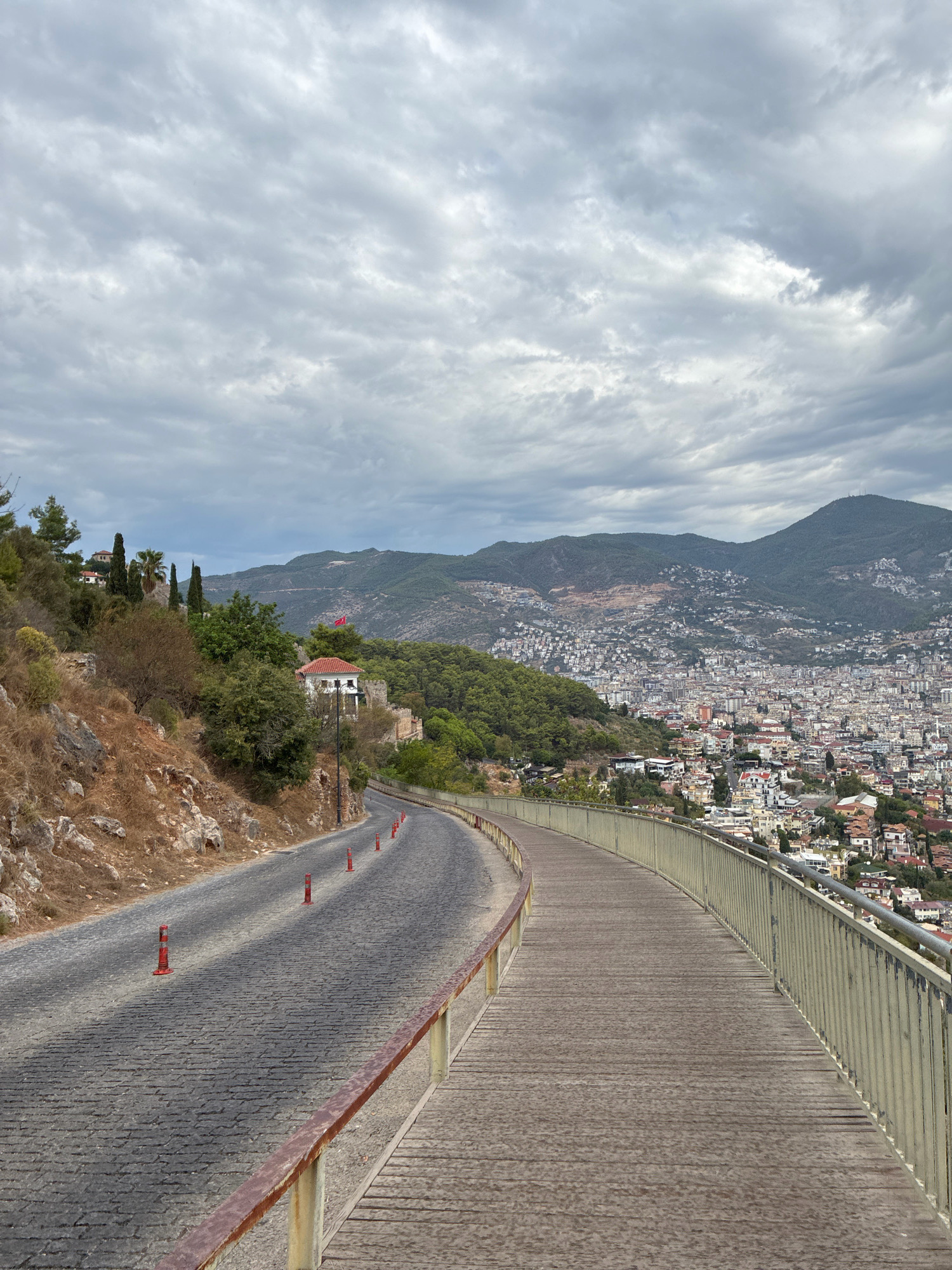 A paved road curves left and uphill to the left of a wooden walkway. The road is lined with orange traffic cones and a rocky, vegetated slope. The walkway is bordered by a metal railing. In the background, a city sprawls across hillsides, leading to a mountain range under a cloudy sky. A red flag is visible atop a white building on the left side of the image. The scene appears to be in Alanya, Turkey.