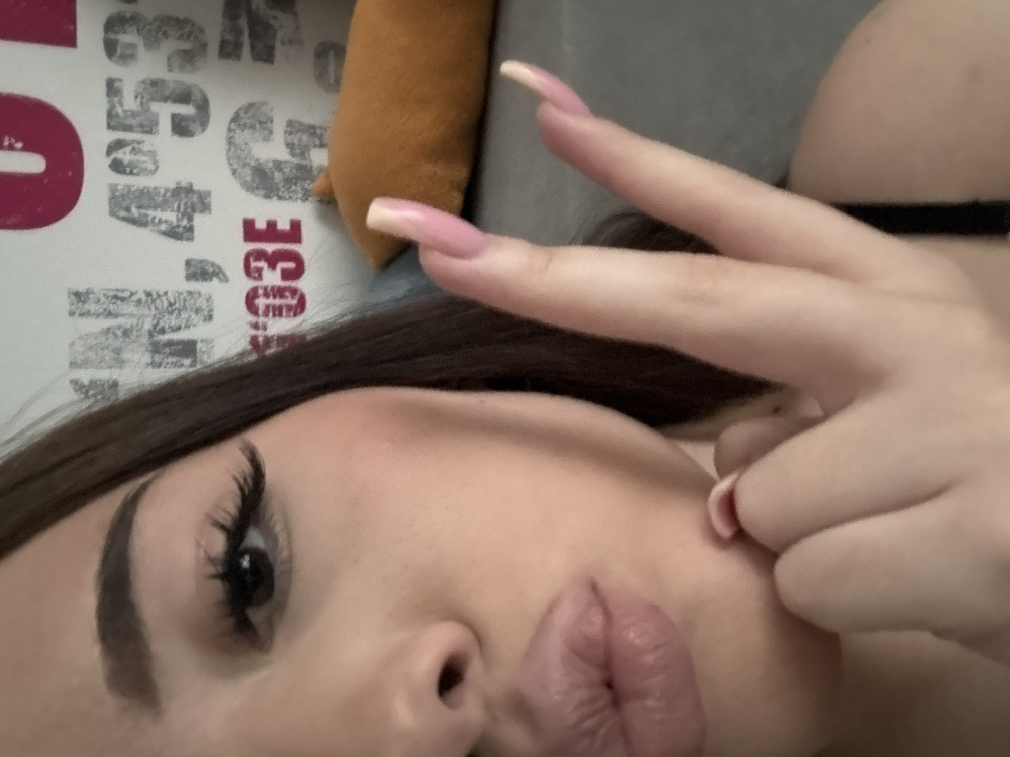 A close-up selfie of a woman's face and hands. Her hair is dark brown, and she has long eyelashes and styled eyebrows. Her lips are slightly puckered. She is holding up two fingers, and her nails are long and pink. The background includes a gray surface, a brown object, and a white patterned material with numbers and letters in red and gray.
It is not possible to determine the location in the city of Naberezhnye Chelny, Russia, from the given image. There are no identifiable landmarks or indicators.