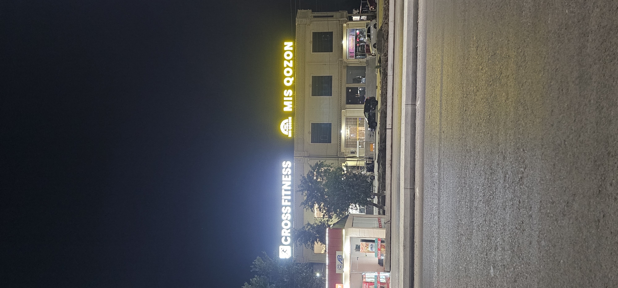 The image shows a building at night in Sheyhancha, Uzbekistan. The building has illuminated signs that read "CROSS FITNESS" and "MIS QOZON". In front of the building, there is a street with parked cars and a small kiosk. The overall lighting suggests it is nighttime, with artificial lights illuminating the building and the street. The environment appears to be an urban or semi-urban area.