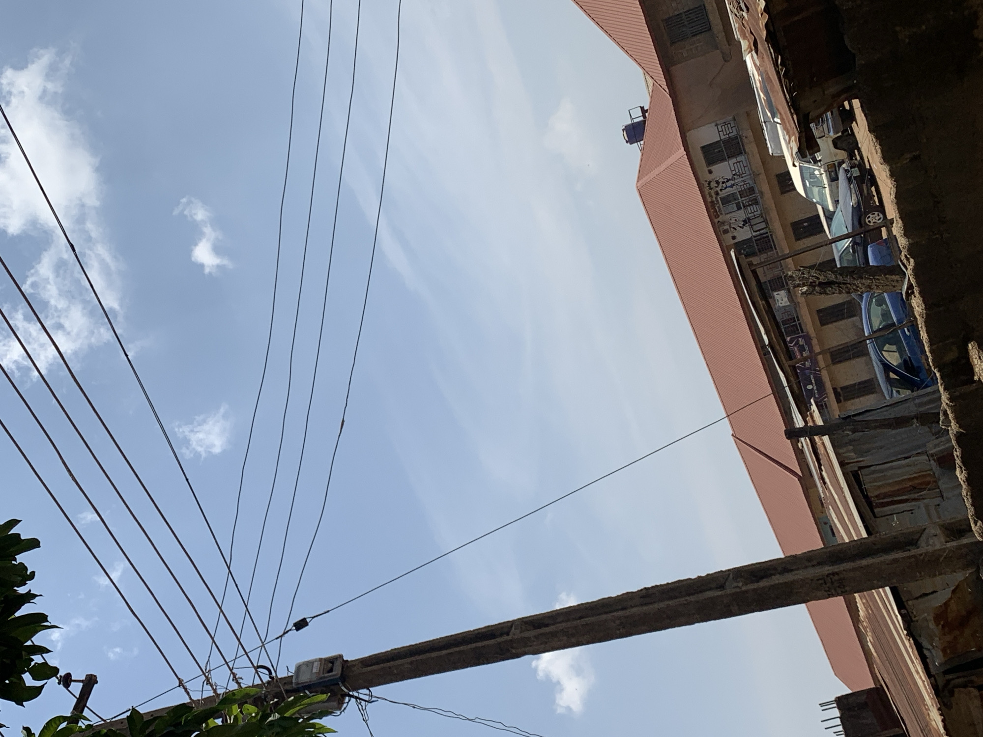 The scene is set in the urban environment of Rafin Par, Nigeria. The viewpoint is tilted upwards, capturing a perspective that emphasizes the urban infrastructure. In the foreground, a weathered concrete utility pole is visible, crisscrossed with numerous electrical wires extending upwards and outwards against a backdrop of a light blue sky, sparsely dotted with small, scattered clouds.
To the right, the frame is filled with a close-up view of a multi-story building. The building appears to be constructed with a mixture of materials, including rusted corrugated iron sheets and concrete. Adjacent to the red roof, there's a small blue water tank. The building has a series of open-air balconies or walkways, where vehicles and parked, specifically a blue car.
The environment suggests an older, possibly lower-income urban area. The sky is bright, indicating daylight, and the electrical wires are a prominent feature of the urban landscape. A few tree branches and leaves are visible in the bottom left corner, partially obscuring the base of the utility pole.