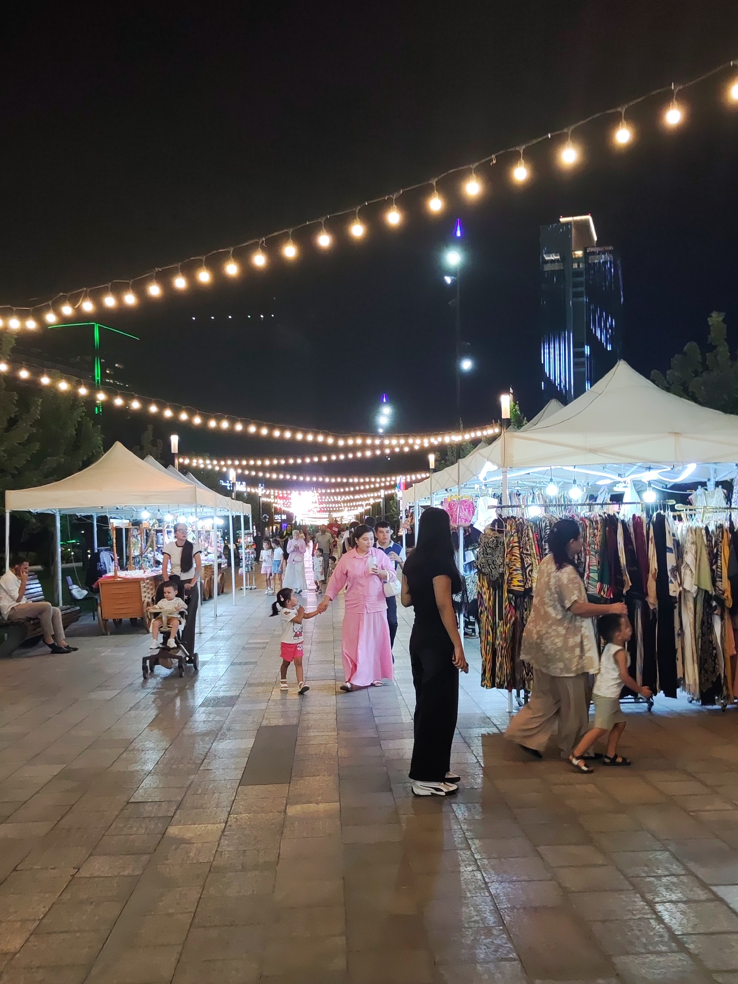 A nighttime scene depicts a bustling outdoor market in Tashkent, Uzbekistan, possibly along Amir Timur Street, characterized by numerous stalls and a lively crowd. The market is illuminated by strings of warm, glowing bulbs suspended overhead, creating a festive atmosphere. Several white canopies, representing market stalls, are visible, displaying a variety of clothing items on racks.
In the foreground, a woman in a black top and pants walks away from the camera, while a young boy in a white shirt and shorts walks beside her. To their left, a woman in a pink outfit holds the hand of a young girl, also in pink, as they walk together. A stroller with a child in it is being pushed by a woman further back in the scene. On the far left, a man in a white shirt and light-colored pants sits on a bench, facing away from the viewer.
In the background, modern buildings, including a tall skyscraper with illuminated blue vertical lines, rise against the dark night sky, suggesting an urban setting. The ground is paved with rectangular stones, and reflections of the lights are visible on the wet surface, indicating recent rain or cleaning. The overall impression is a vibrant, active marketplace under the night sky.