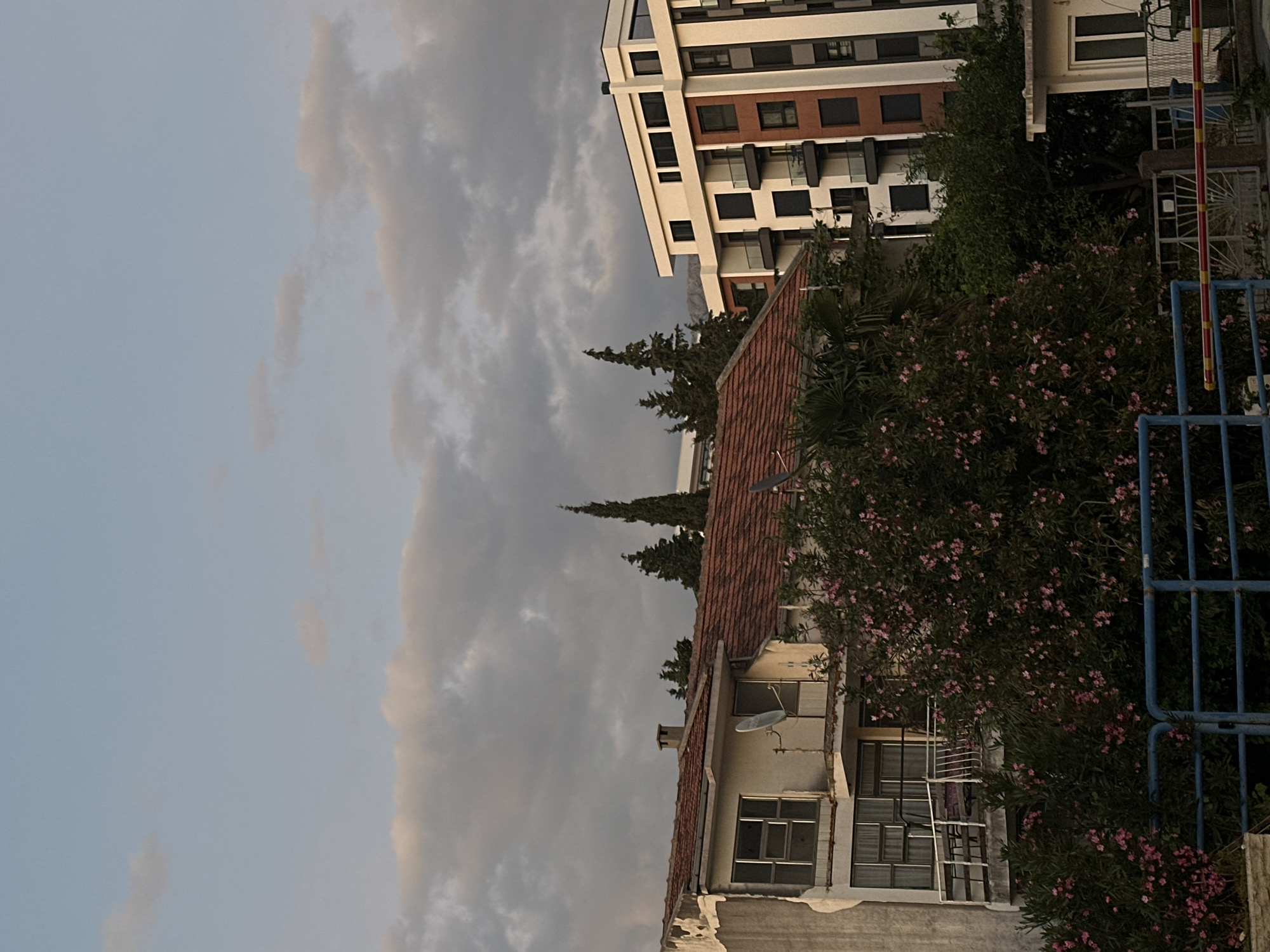 The image shows a view in Šušanj, Montenegro, featuring a combination of urban and natural elements. In the foreground, there's a residential building with a red tile roof, partially obscured by lush green vegetation with pink flowers. A satellite dish is mounted on the side of the building. Adjacent to this, a modern apartment building with a white facade and brown accents is visible. The background is dominated by a cloudy sky, with tall evergreen trees interspersed between the buildings. A blue metal fence is also partially visible. The overall scene suggests a residential area with a blend of older and newer architecture, situated within a landscape with substantial tree cover.