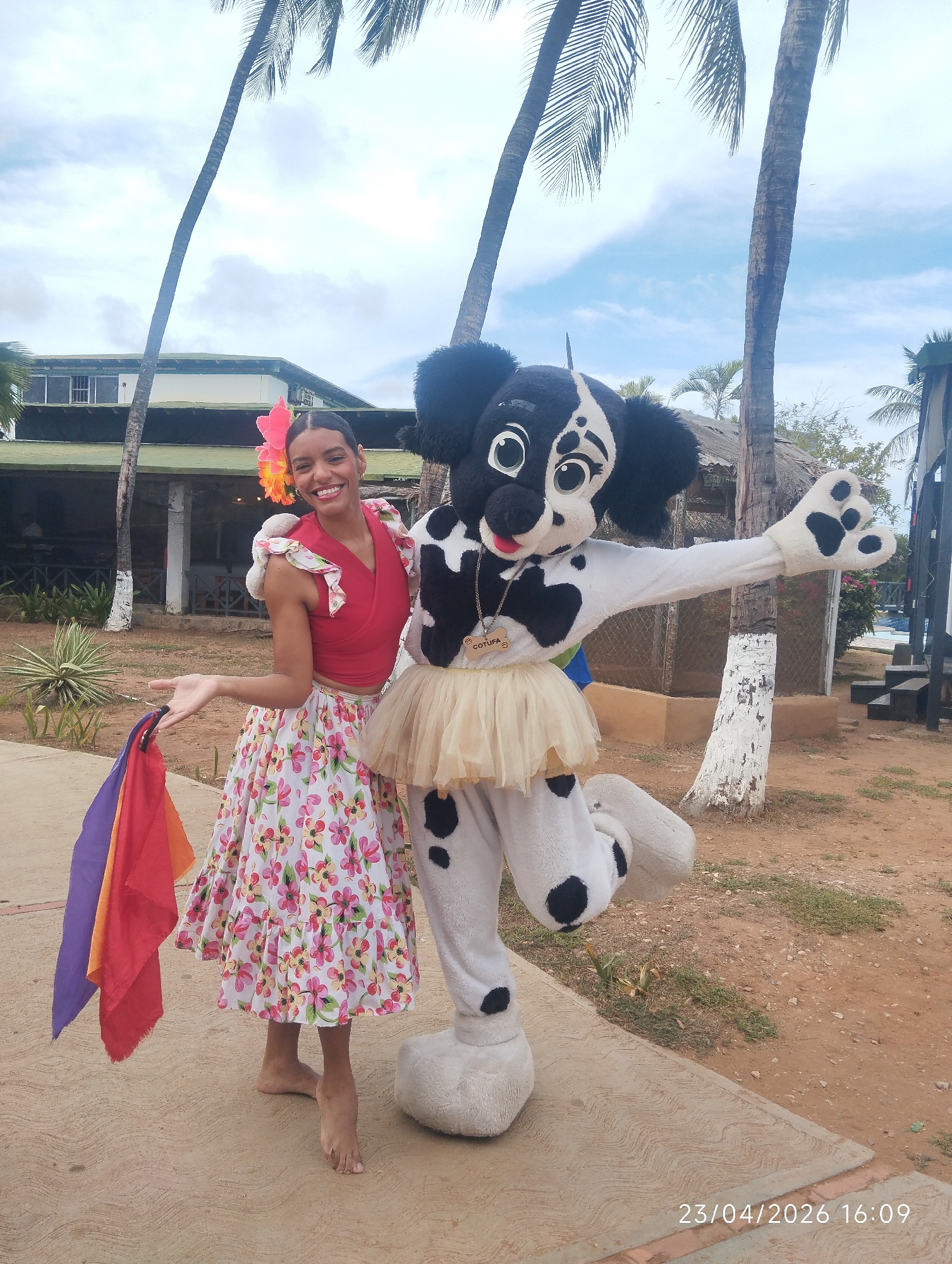 A smiling woman and a person in a dog mascot costume pose cheerfully for a photo outdoors. The woman, positioned on the left, has dark hair decorated with large pink and orange flowers. She wears a red short-sleeved top with ruffled details and a long, flowing white skirt patterned with pink, yellow, and green flowers. Barefoot, she holds a bundle of colorful scarves (purple, red, and orange) in her left hand and gestures with her right, displaying a wide smile.
Beside her, the mascot, a black and white spotted dog with large, expressive cartoon eyes, a black nose, and a visible red tongue, stands in a dynamic pose with one leg raised and an arm outstretched. It wears a light beige tutu around its waist, and a bone-shaped tag with the text "COTUFÁ" hangs from its neck.
The background features several tall palm trees reaching towards a partly cloudy sky. Behind the subjects, various buildings are visible, including a structure with a green roof and white pillars, and another with a thatched roof, suggesting a resort or tropical setting. The ground is a mix of concrete paving and dirt, with some sparse vegetation. The scene is brightly lit, consistent with late afternoon, as indicated by the timestamp "23/04/2026 16:09" in the bottom right corner. The overall atmosphere is joyful and lively.