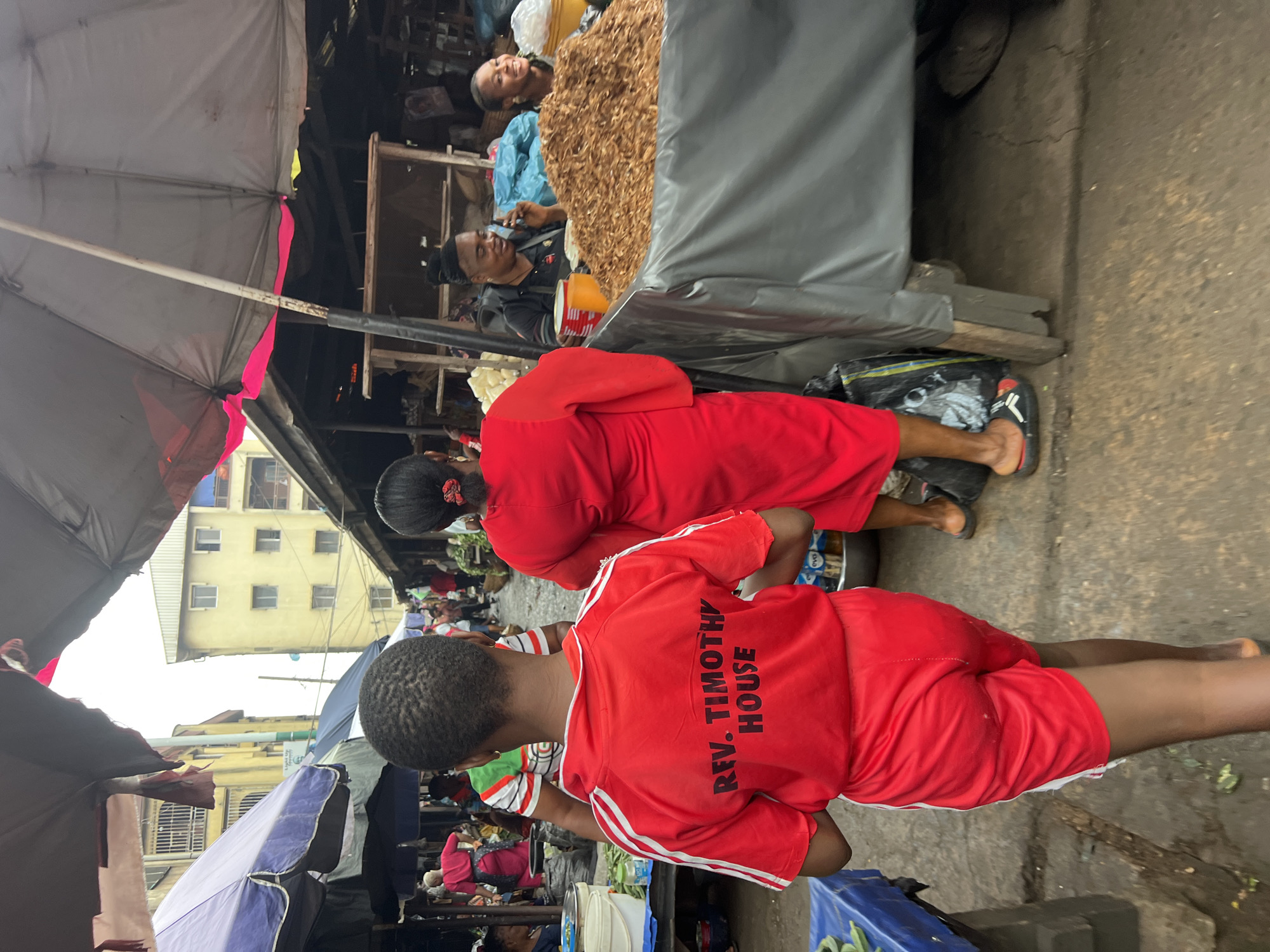 The image depicts a busy outdoor market scene in Nkpor, Nigeria. In the foreground, a young person wearing a red shirt with "REV. TIMOTHY HOUSE" printed on the back and matching red shorts walks away from the camera. To their right, an adult wearing a bright red dress walks in the same direction.
The market is characterized by various stalls covered by umbrellas and tarps. To the left, a blue tarp covers a display of green produce, possibly plantains. In the background, several buildings are visible, including a tall, light yellow building with multiple floors and windows.
Other people are present, including two individuals at a stall to the right. One person, with dark hair pulled back, is speaking on a mobile phone and smiles. Beside them, another person smiles broadly while looking towards the camera. A large mound of dried shrimp or similar small seafood is displayed on the counter of this stall.
The ground appears to be a mixture of concrete and dirt, and the overall lighting suggests an overcast or early morning/late afternoon setting. The atmosphere is that of a typical bustling Nigerian market with a variety of goods and people engaged in commerce.