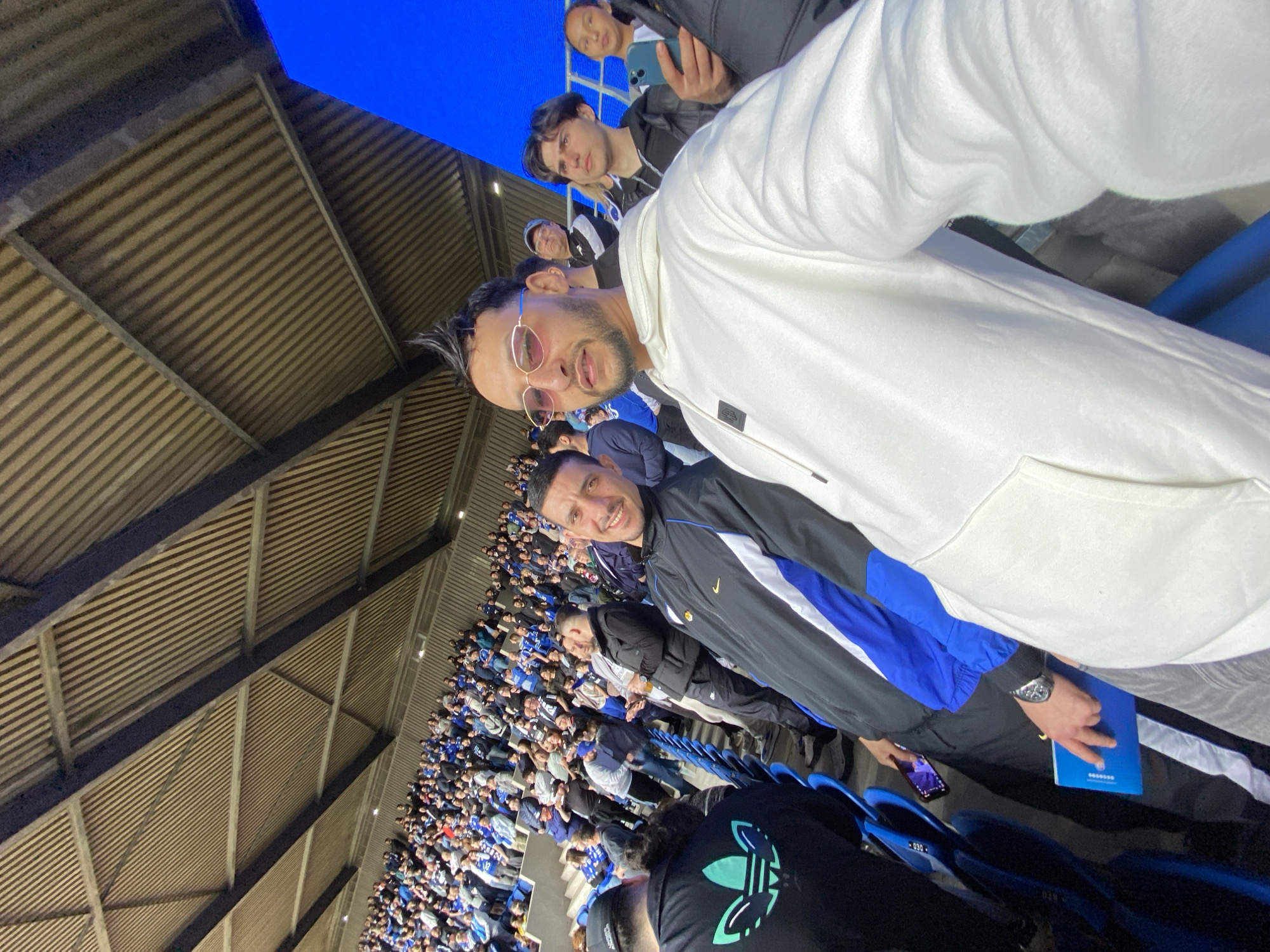 A selfie-style photograph depicts a crowd of people in an open-air stadium. The stadium features a roof with horizontal ridges. Many people in the crowd are wearing blue or white clothing. The primary subject is a man with a beard wearing sunglasses and a white hoodie. Behind him, other people are visible, including one taking a picture with a smartphone. The image is taken at an angle, looking down into the crowd. The setting appears to be in the city of Strasbourg, France, specifically at a stadium likely hosting a sporting event.