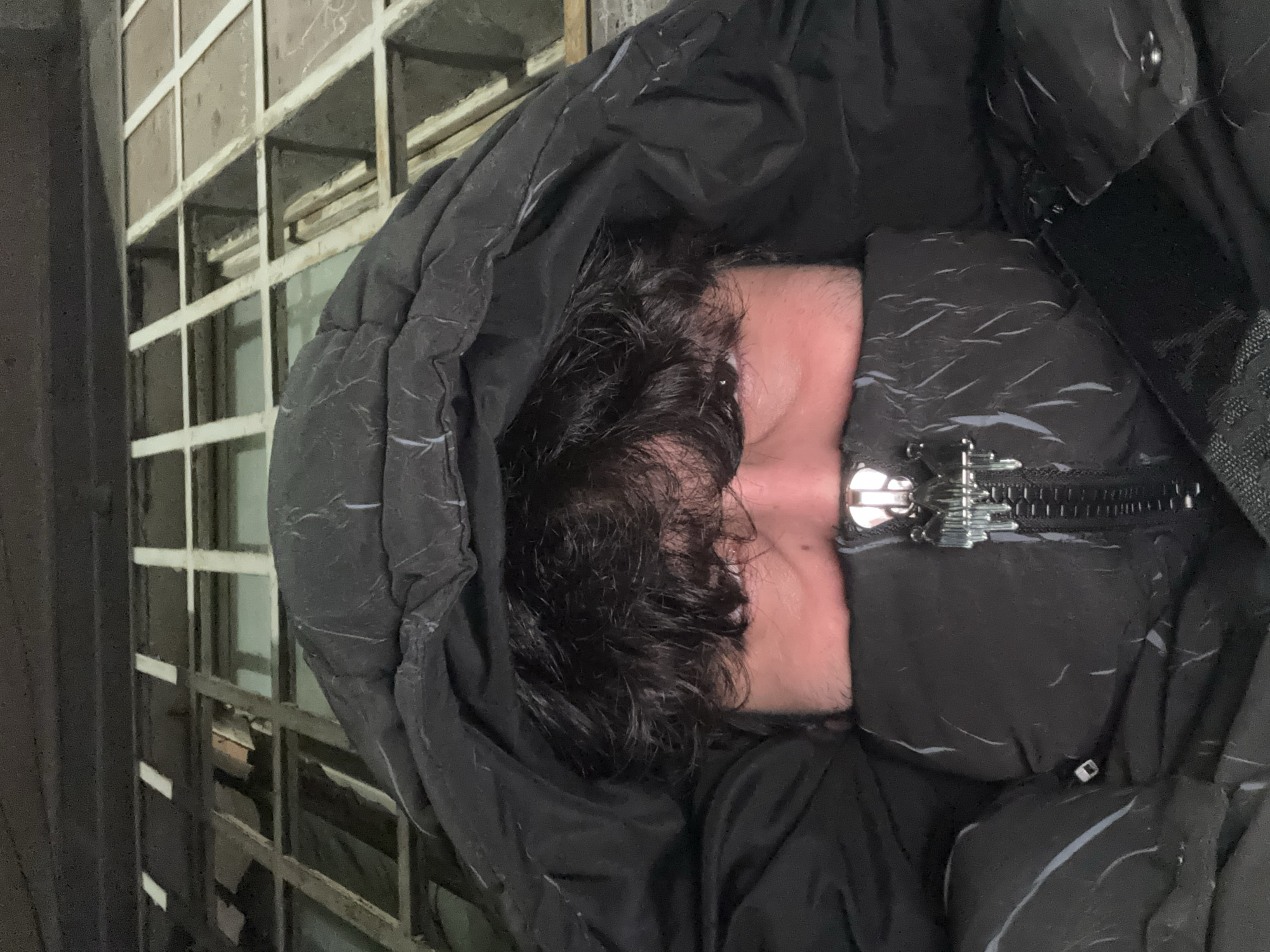 A person with dark, curly hair is almost entirely concealed within a dark-colored, hooded puffer jacket. The jacket is zipped partially up, and the zipper's metal pull is visible. Only a portion of the person's face, appearing pinkish, is visible between the hair and the jacket. The background shows a metal-framed structure with a grid-like pattern. Based on the aesthetic and potential context clues of a "down jacket" it is possible it is cold season in Yerevan, Armenia, if that is where the photo was taken. There is no further information to ascertain its location in Yerevan.