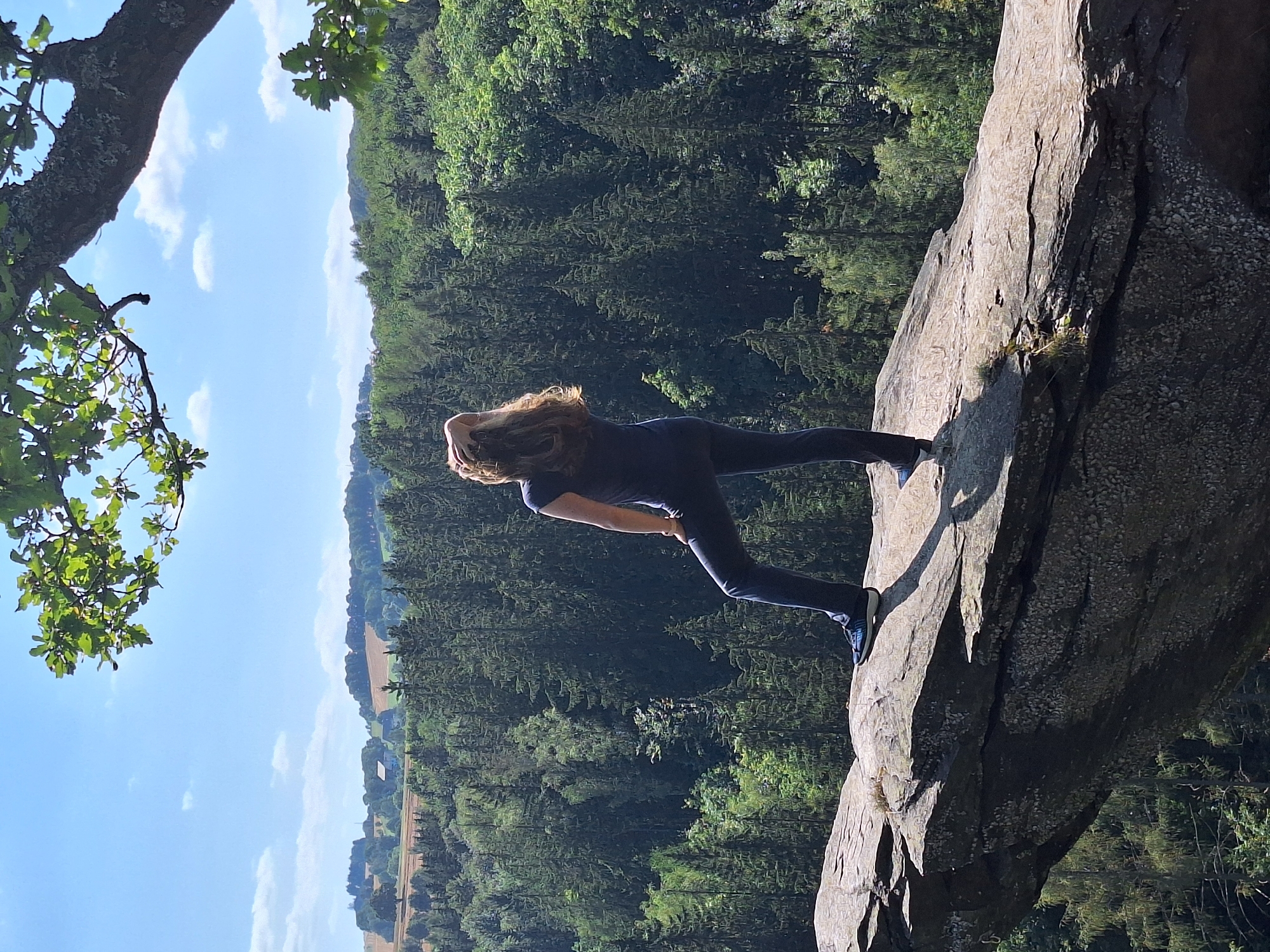 A woman with long, brown hair stands on a rocky outcrop, her back to the camera, looking out over a dense forest and a rolling, rural landscape under a blue sky with scattered clouds. She is wearing dark blue leggings and a dark blue t-shirt, and her right hand is resting on the back of her head. Her left leg is bent at the knee and extended slightly back. The rocky outcrop she is standing on is textured and appears to jut out over the forested valley. In the distance, fields and a few buildings are visible on the hills. The scene is framed by a large, moss-covered tree branch with green leaves in the upper right corner of the image. The location is likely the "Piscatorfelsen" (Fisherman's Rock) in the Wolkenstein, Germany area, known for its dramatic rock formations and forest views.