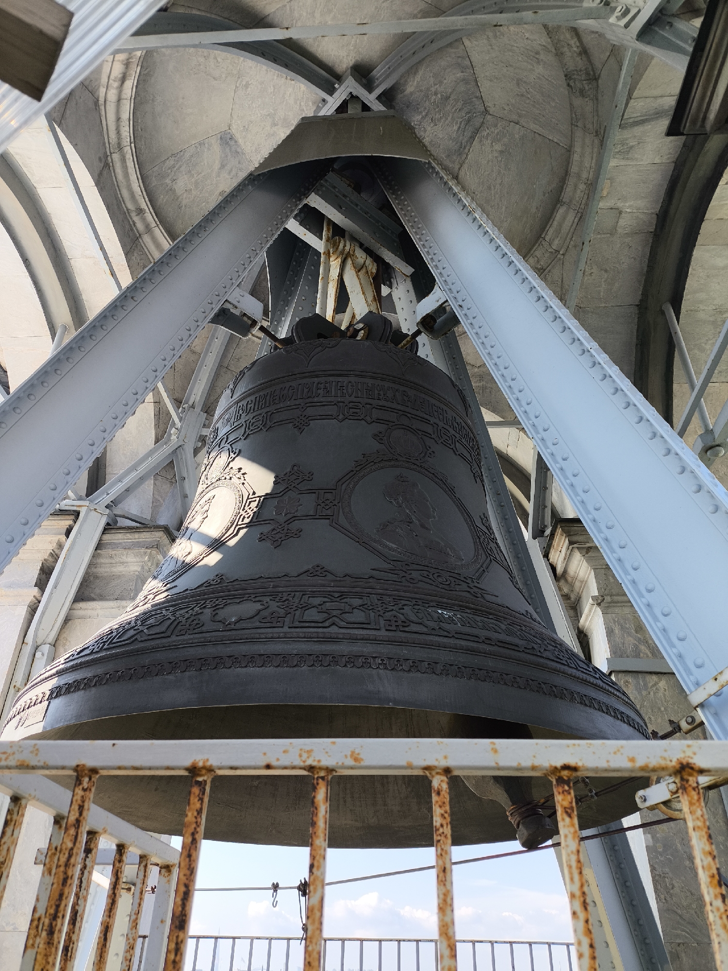 A low-angle, outdoor, close-up shot captures the interior of a bell tower in Saint Petersburg, Russia, focusing on a large, dark bronze bell suspended from a metal structure. The bell, adorned with intricate relief carvings, is the central subject. The metallic beams supporting the bell are painted a pale blue and secured with numerous rivets. Above the bell, a stone dome with arched architectural elements is visible, constructed from large, grey stone blocks. In the foreground, a rusted, white metal railing with vertical bars suggests a viewing platform. The background shows a bright, partly cloudy sky. The lighting suggests it is daytime.
