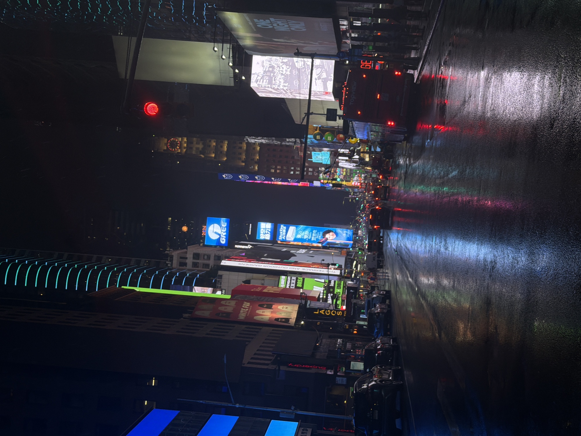The image depicts a nighttime scene in Times Square, New York City, following a rain shower. The wet asphalt street reflects the vibrant neon lights of towering billboards and buildings. Several cars are parked and in motion on the street, with their headlights and taillights illuminated. A red tour bus is visible in the foreground, displaying "TopView" on its rear. Pedestrians are seen on the sidewalk. The overall atmosphere is lively and bustling, characteristic of this iconic urban location.