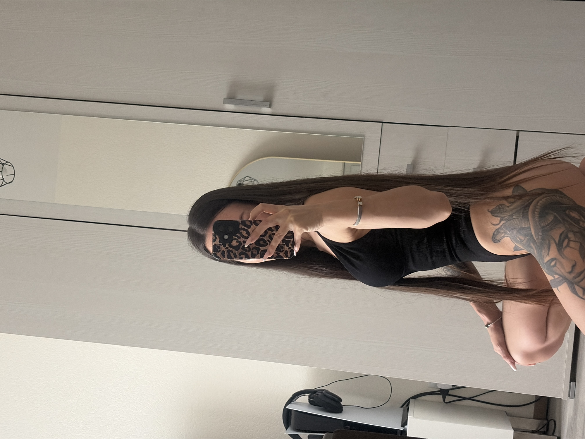 A woman with long, dark hair is photographed indoors against a light-colored wall and furniture. She is partially visible, wearing a black top, with a significant tattoo on her shoulder and upper arm. She is holding a leopard-print phone up to take a picture of herself in a mirror on what appears to be a wardrobe. A desk is below the wardrobe, featuring a gaming console and headset. The specific location within the city of Unknown, Unknown, is indiscernible from the image.