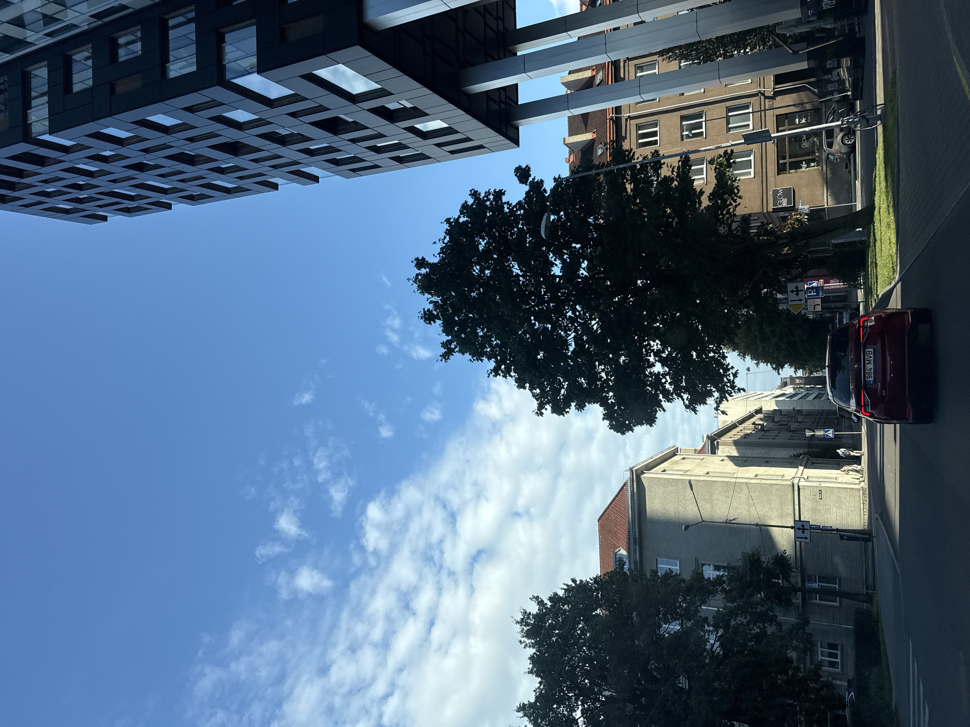 The image shows a view from a vehicle driving on a street in Tallinn, Estonia. On the right is a modern, black, multi-story building with numerous square windows, supported by thick concrete pillars. To the left of this building, a darker, older, multi-story building with a reddish-brown roof is visible. Further left, another light-colored, multi-story building with multiple windows is seen. A large, dark green tree with dense foliage stands in the foreground, partially obscuring the view of the buildings behind it. In the street, a red car with license plate 984 MVB is traveling away from the camera. Two pedestrians are walking on the sidewalk to the left of the street, near a traffic sign with directional arrows. The sky is blue with scattered white clouds. The street is paved with asphalt, and there is a grassy median on the right side of the road. The scene appears to be a city street during the daytime.