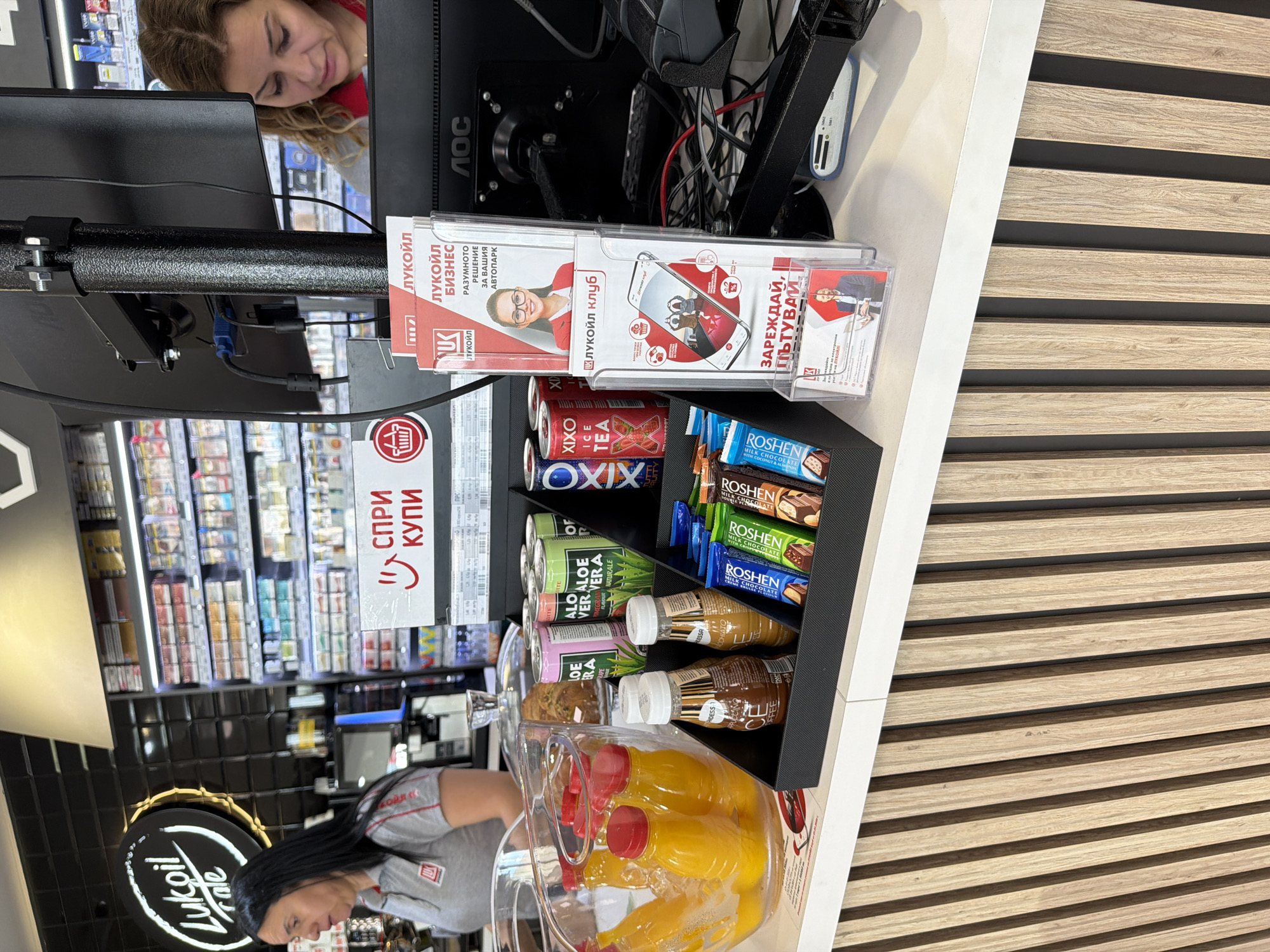 The image captures a scene inside a Lukoil service station in Chuchuligovo, Bulgaria. In the foreground, a counter displays a variety of beverages and snacks. To the left, a glass basin holds several small plastic bottles filled with a yellow liquid, likely juice, submerged in ice. Adjacent to this are several cans of Aloe Vera drinks, some labeled "Aloe Vera Original" and "Aloe Vera Pomegranate". Further to the right, a black display organizer holds an assortment of chocolate bars, including "Roshen Milk Chocolate", "Roshen Chocolate", and other unidentified brands. Behind the counter, a computer monitor is visible. To the left of the monitor, a woman with dark hair is standing, wearing a grey Lukoil branded shirt. To the right of the monitor, a woman with curly blonde hair is seated and looking down. Several promotional brochures and leaflets are displayed on a stand in front of the monitor. The overall environment suggests a retail setting within a gas station, focusing on impulse purchase items.