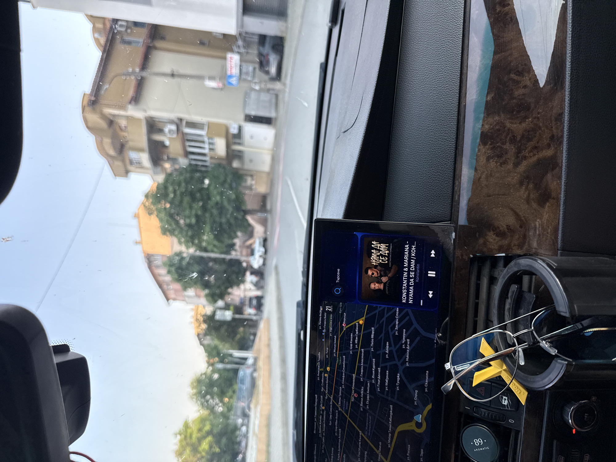 The image presents a view from inside a vehicle, looking forward through the windshield onto a city street in Stara Zagora, Bulgaria.
The interior of the car features a black dashboard with a subtle crack or seam visible above the passenger side. A central infotainment screen displays a navigation map on the left, showing street names in Cyrillic, consistent with Stara Zagora. The map indicates the vehicle's approximate location near the intersection of ul. Vasil Levski and ul. Georgi Apostolov. The right side of the screen displays a music player interface, showing "KONSTANTIN & MARIANA" performing "NYAMA DA SE DAM / KOH...", with an accompanying album cover featuring two individuals. Below the screen, in a black cup holder, a pair of silver-framed eyeglasses with blue lenses rests, along with a yellow plastic item marked "Aromatic". To the left of the cup holder, climate controls are visible, including a circular "OFF" knob and a digital display showing "89°". The windshield exhibits minor smudges and dust. A dark, angular object, possibly a dash camera or rearview mirror housing, is mounted at the top center of the windshield.
The exterior scene, viewed through the windshield, depicts a sunny daytime environment. Multi-story buildings, predominantly yellowish-beige with red-tiled roofs, line both sides of the street. One building on the right features multiple balconies and visible air conditioning units. Abundant green trees are present along the road, suggesting warm weather. A pedestrian crossing sign (blue triangle with white pedestrian symbol) is partially visible on the right sidewalk, alongside a large grey rectangular trash bin and a street light pole. Several other vehicles, including a dark car on the left and a silver/grey car on the right, are visible further down the street. The sky is clear and light blue. No individuals are visible in the scene, either inside or outside the vehicle.
