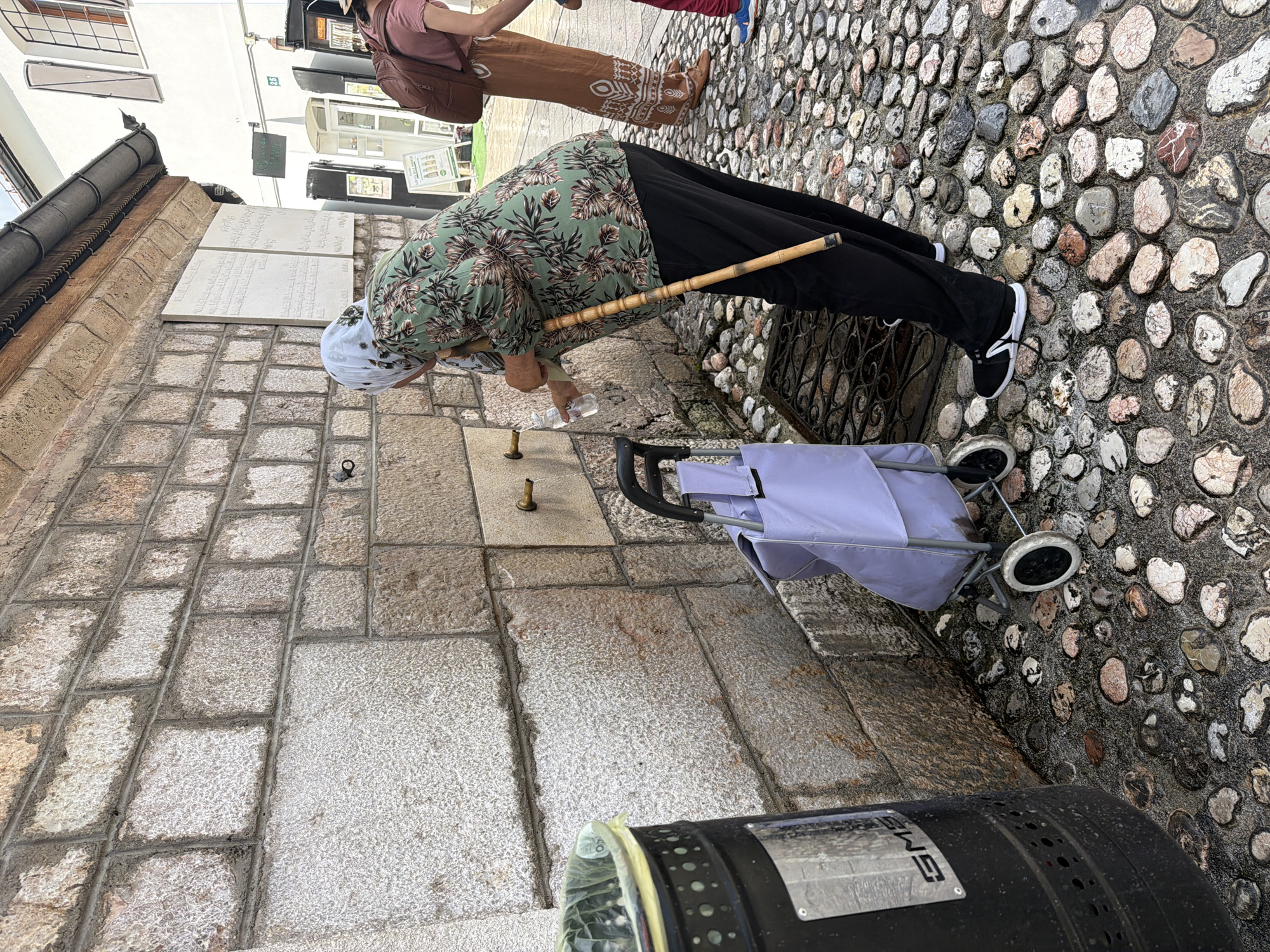 In Sarajevo, Bosnia and Herzegovina, a woman wearing a headscarf and a floral-patterned shirt is using a public fountain. She is holding a plastic bottle under one of the spigots, collecting water. Beside her stands a wooden walking stick. In front of the woman is a light purple shopping trolley with two wheels. To the left of the scene, a large black trash bin with a yellow liner is visible. The ground is paved with numerous cobblestones of varying colors and sizes. Behind the woman, a stone wall is prominent, with two decorative inscribed panels mounted on it. Another woman, wearing a brown patterned skirt and a pink top, with a brown backpack, is partially visible to the right. The overall setting appears to be an outdoor public area with architectural elements consistent with historic European cities.